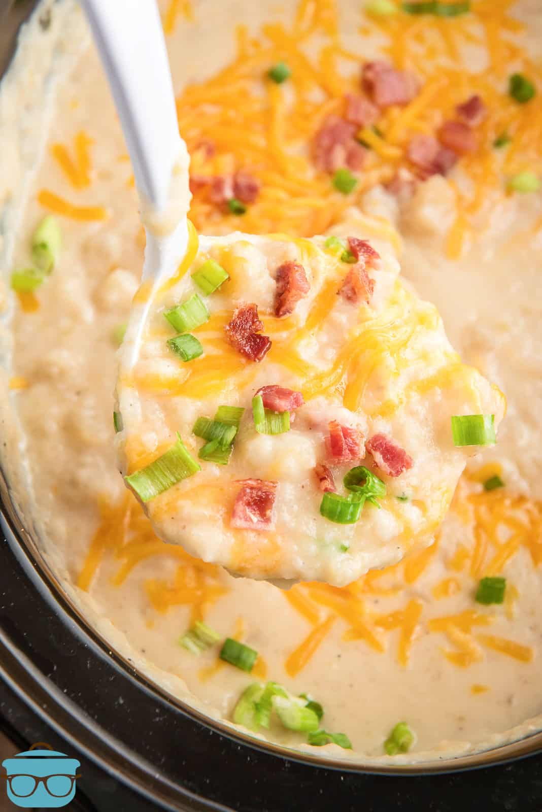 a large ladle of potato soup with cheese and bacon being held over a slow cooker full of potato soup.