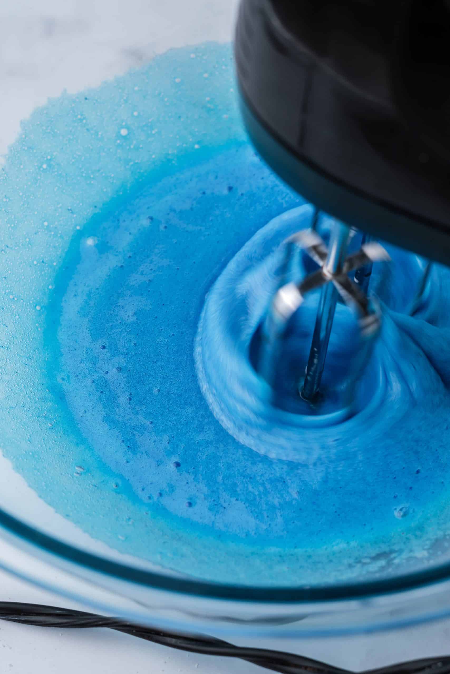 using a handheld electric mixture to mix blue egg whites in a clear bowl.
