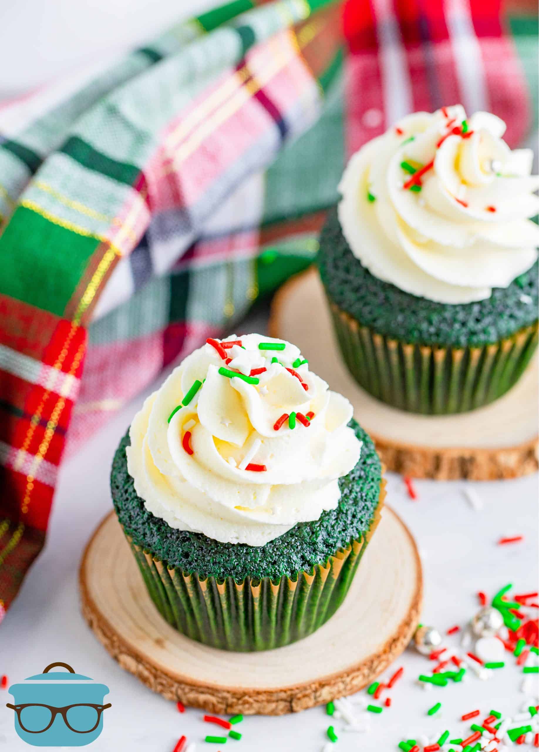 two green velvet cupcakes with buttercream frosting displayed on two small circular wooden discs with a plaid dish towel in the background.