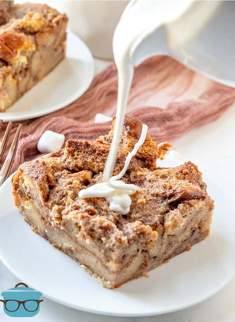 pouring marshmallow cream sauce from a white jar on top of a slice of bread pudding that is on a small white plate.
