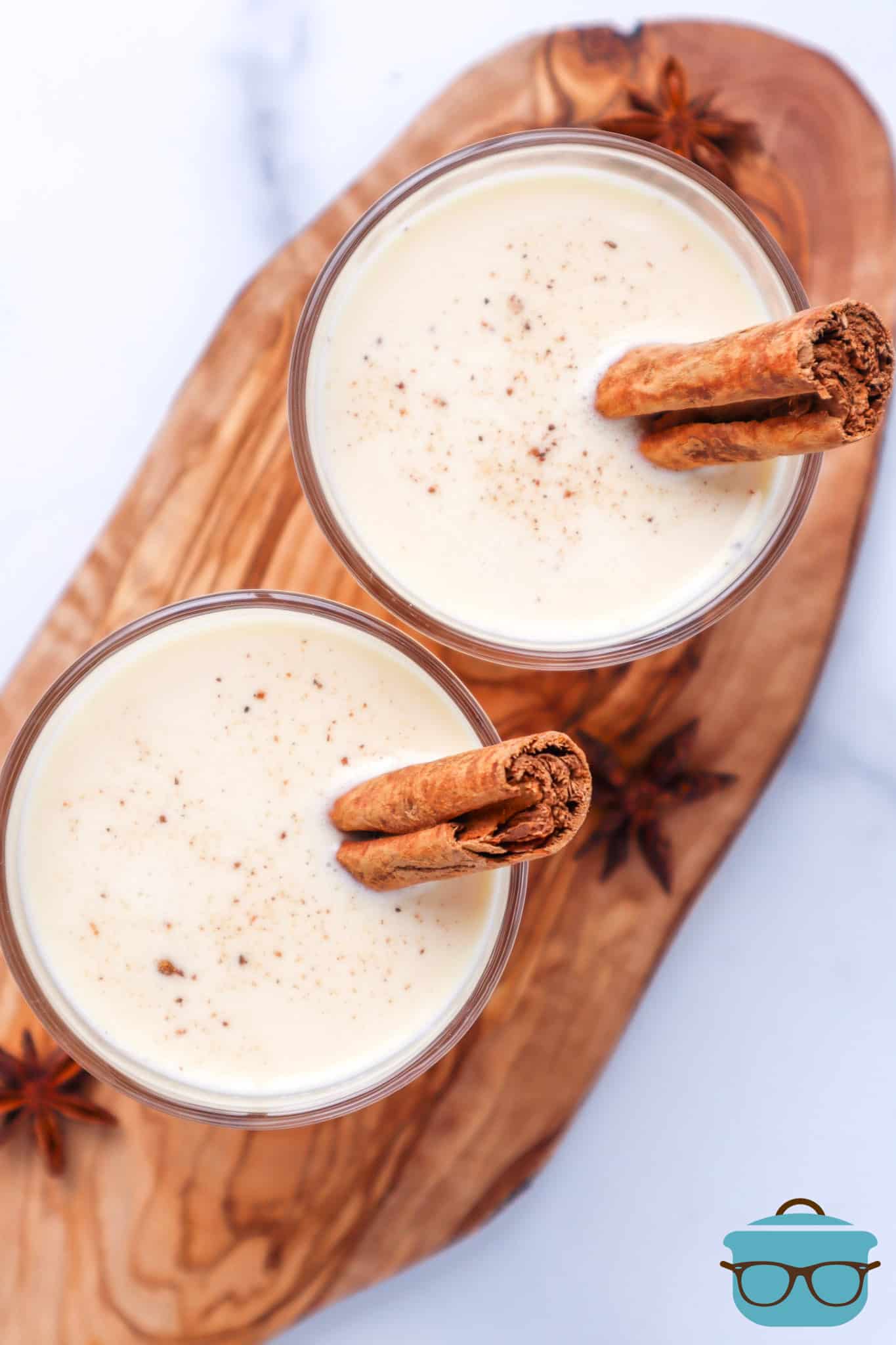 Instant Pot Egg Nog shown in two small, clear glasses with sticks of cinnamon in each glass shown on a wooden cutting board.