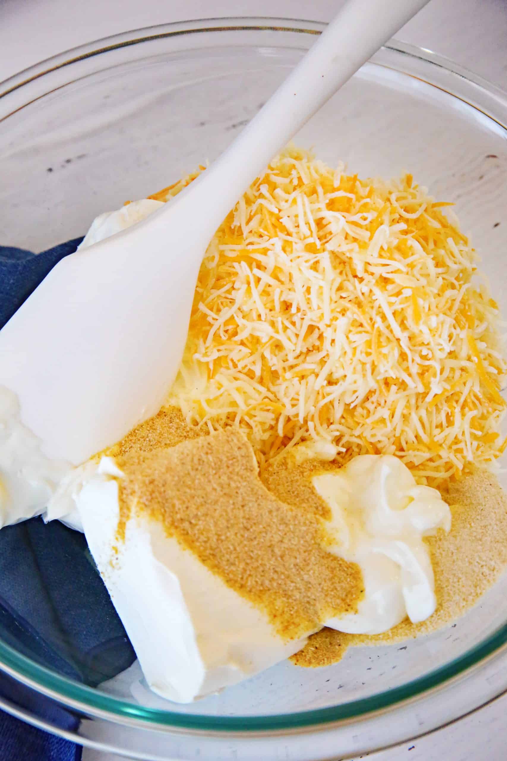cream cheese, shredded cheese, mayonnaise, garlic powder, and onion powder in a large clear bowl with a white spoon.