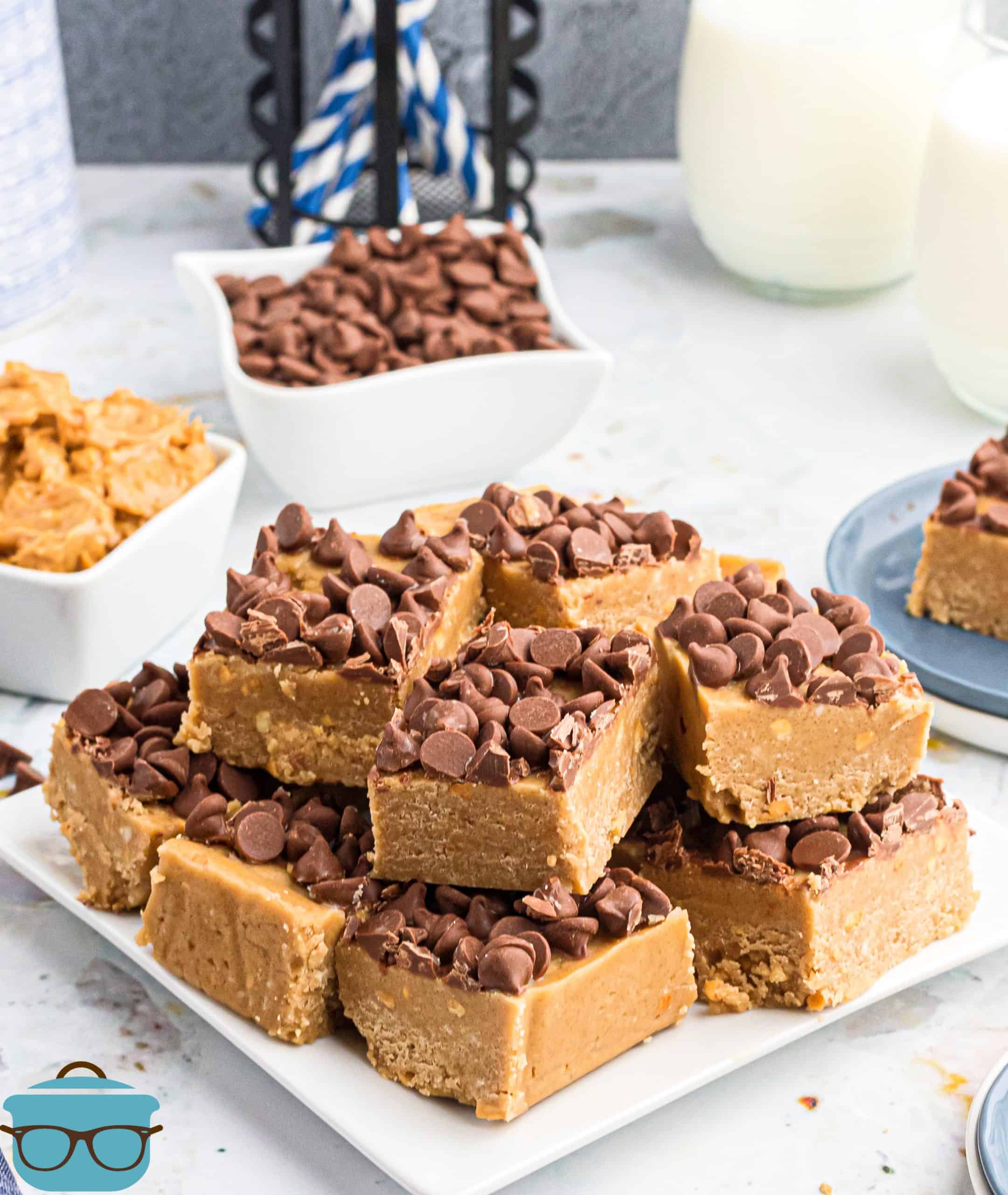 pieces of peanut butter fudge topped with chocolate chips shown on a white square plate with bowls of chocolate chips and peanut butter in the background.