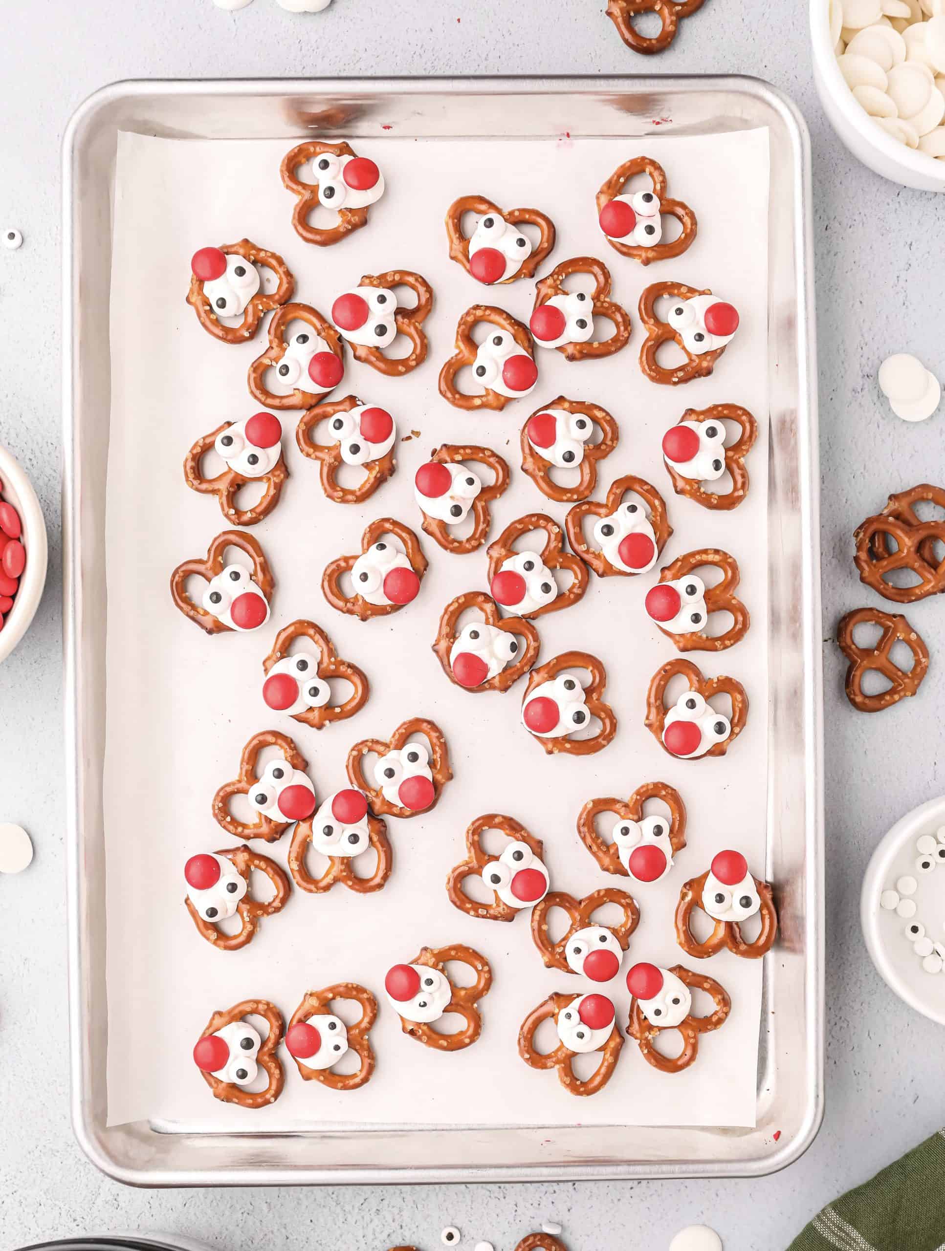 mini pretzels in a single layer with white chocolate dots and red m&m chocolate candies to make a reindeer looking pretzel on a baking sheet.
