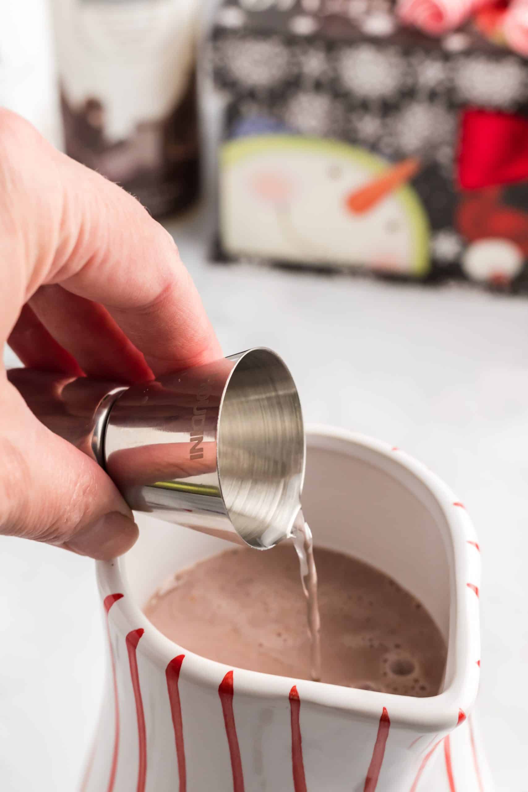 adding a jigger of whipped cream flavored vodka to hot cocoa mix in a white mug.