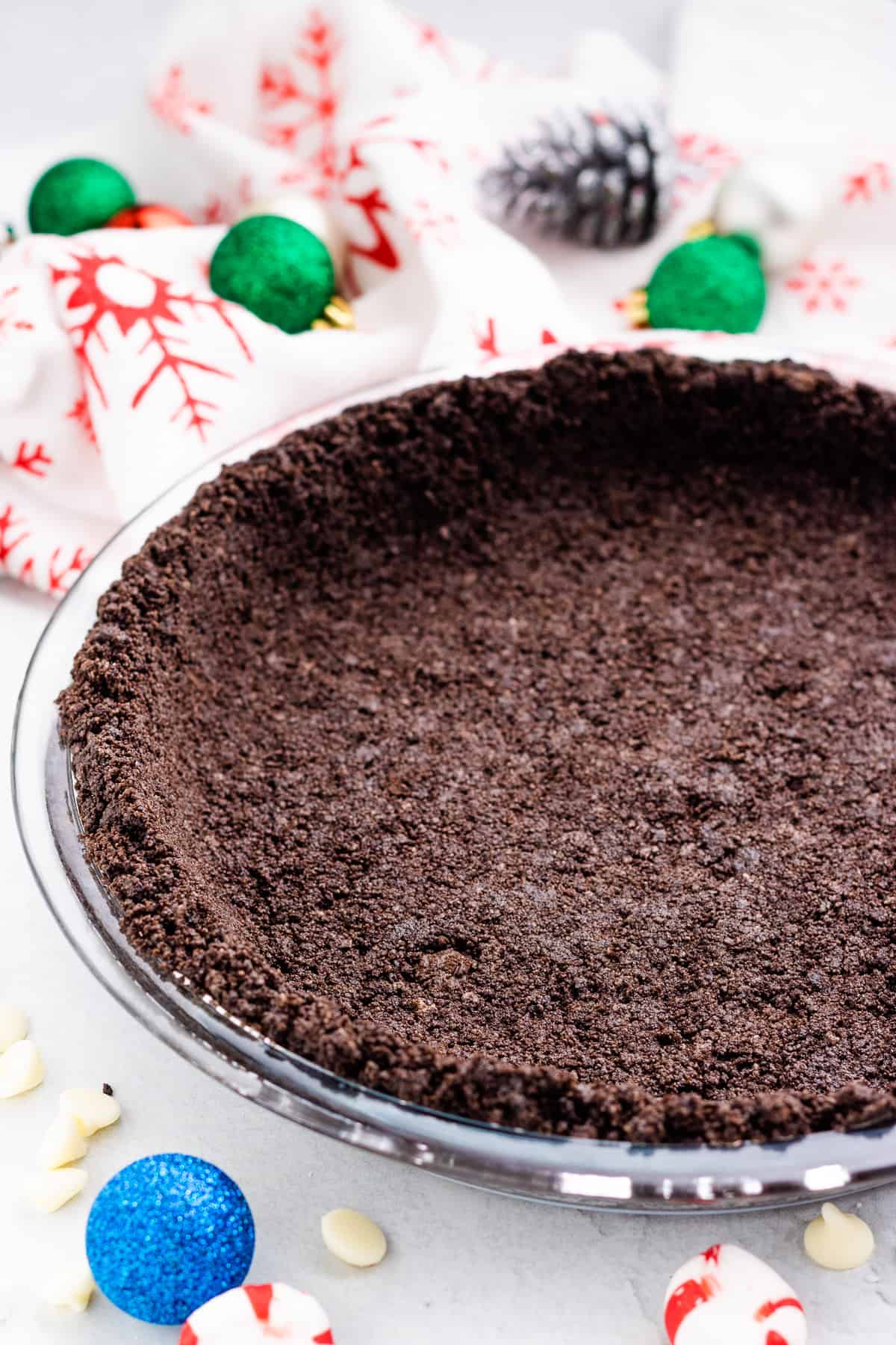 Oreo Cookie pie crust in a clear round pie pan with small Christmas decorations in the background.
