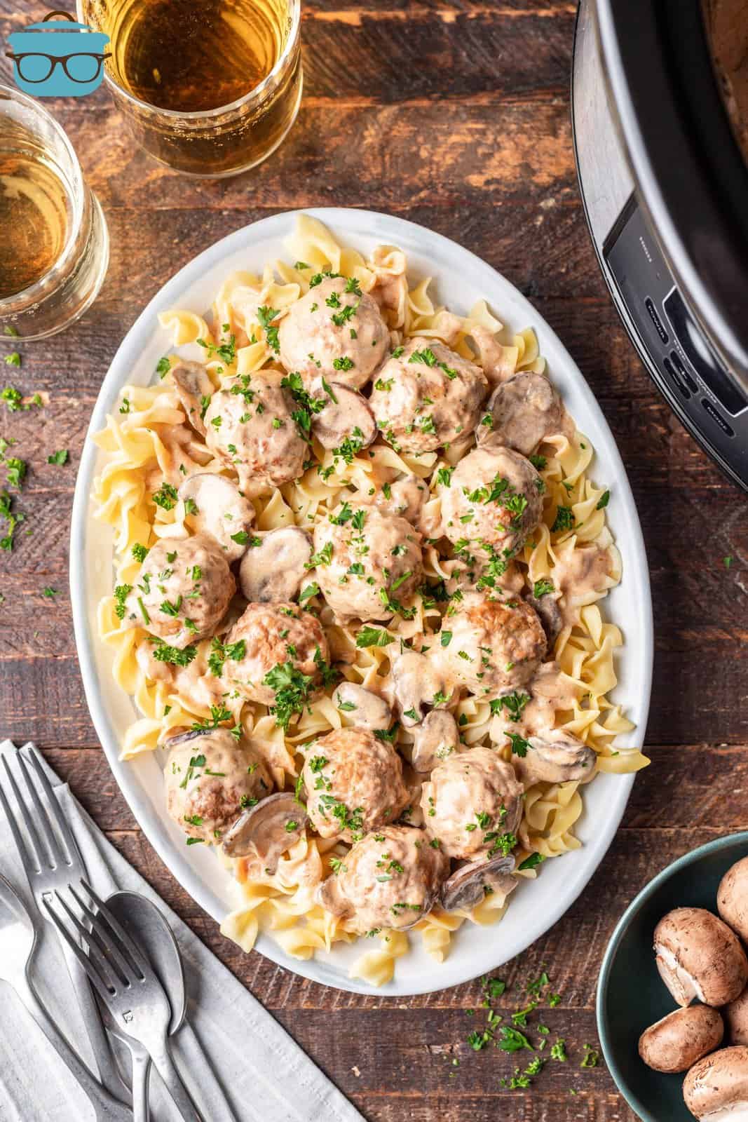 an overhead photo of egg noodles topped with Crock Pot Meatball Stroganoff on a large oval plate on a wooden surface with a slow cooker sitting off to the side. 