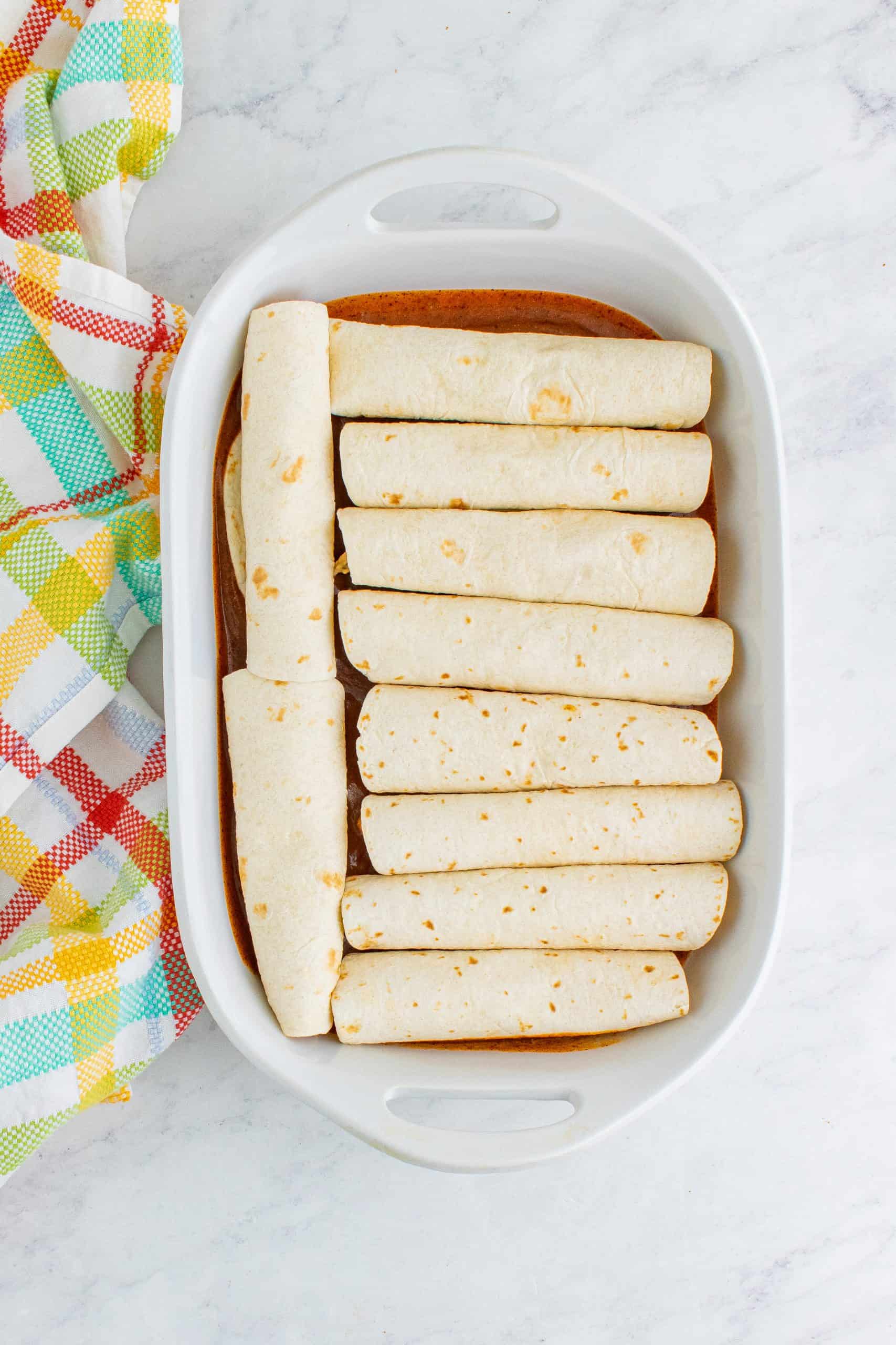 10 rolled tortillas placed in a single layer in a white baking dish.
