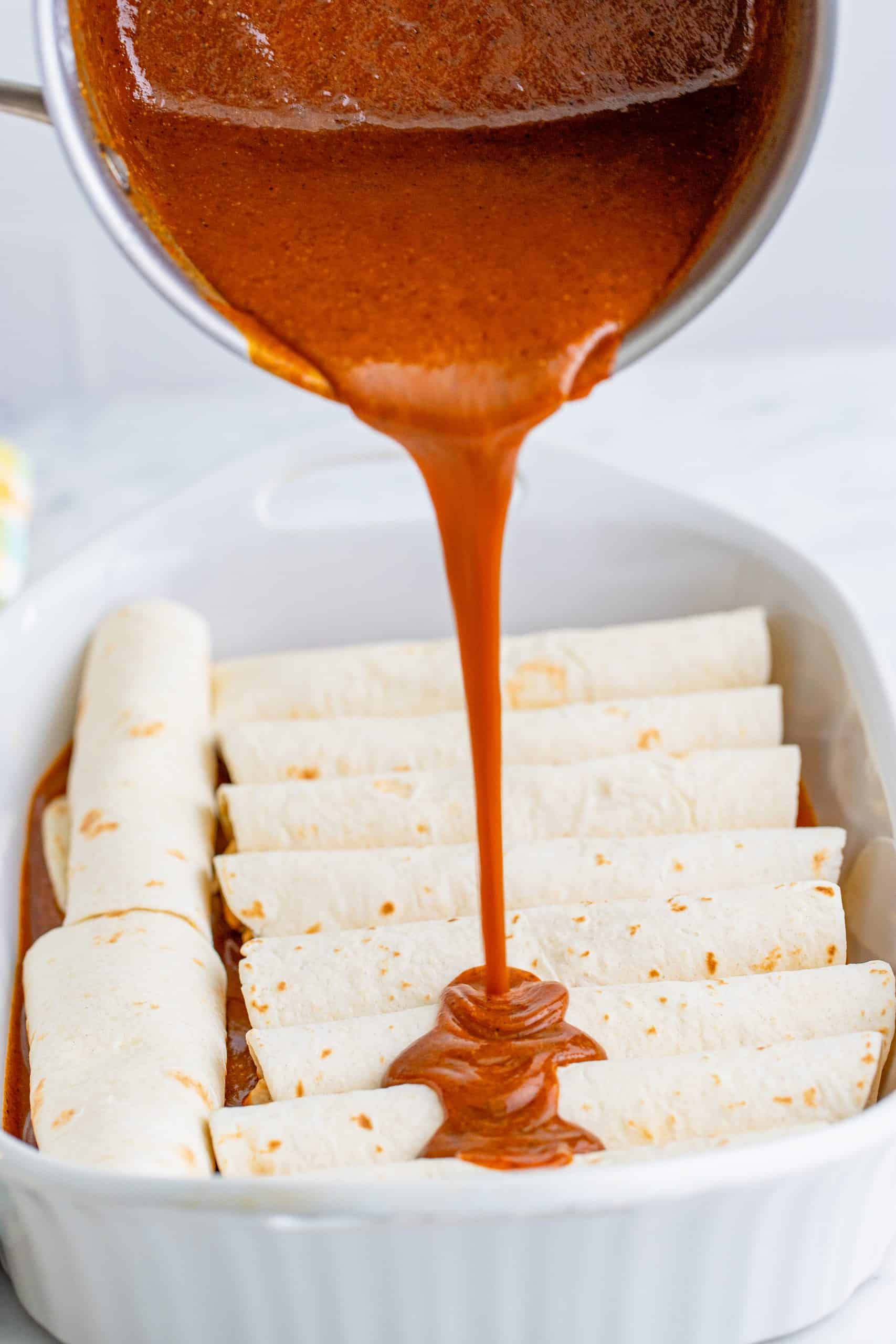 pouring enchilada sauce on top of rolled flour chicken enchiladas in a baking dish.