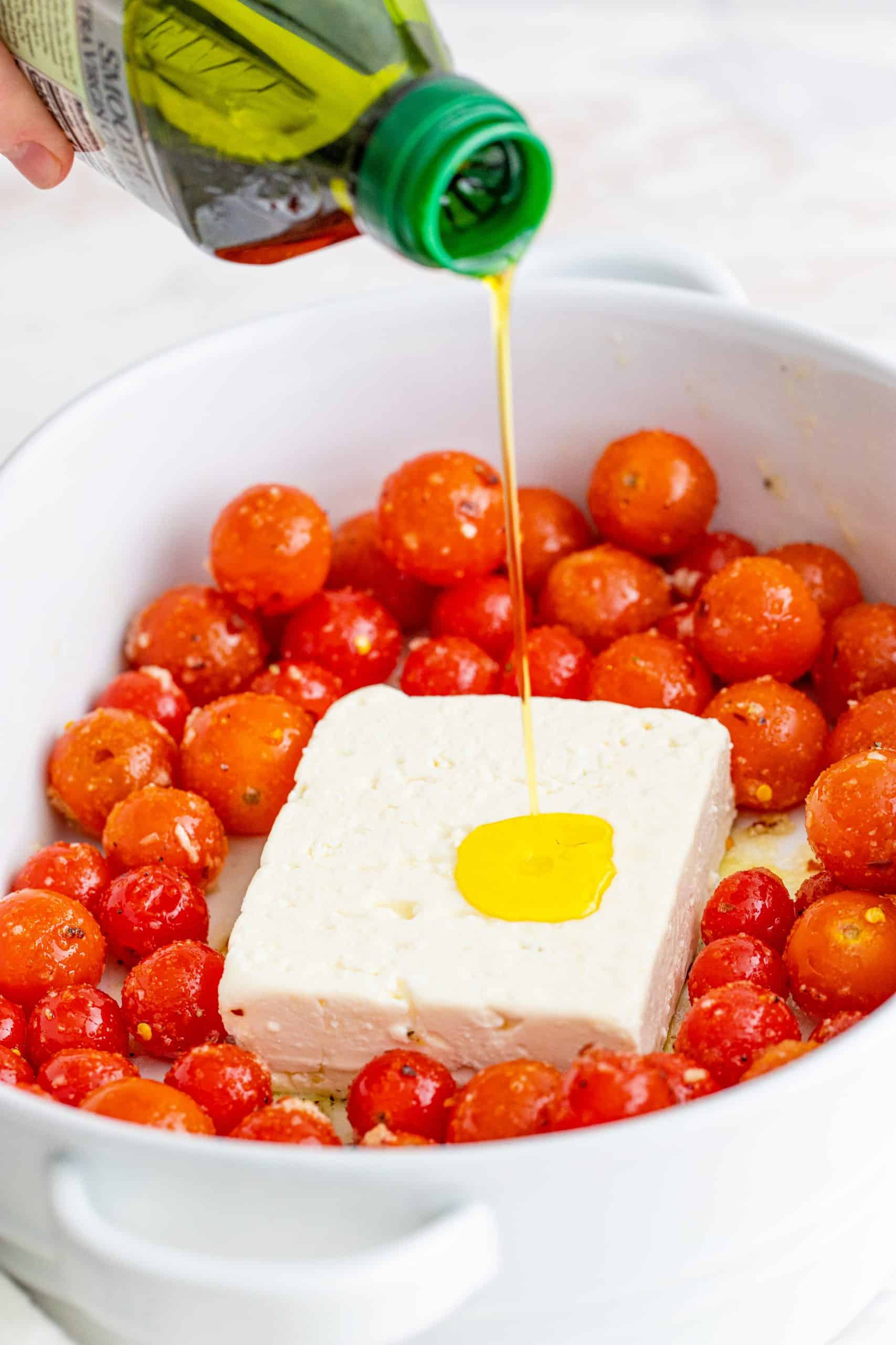 pouring olive oil on top of block of feta cheese and tomatoes in baking dish.
