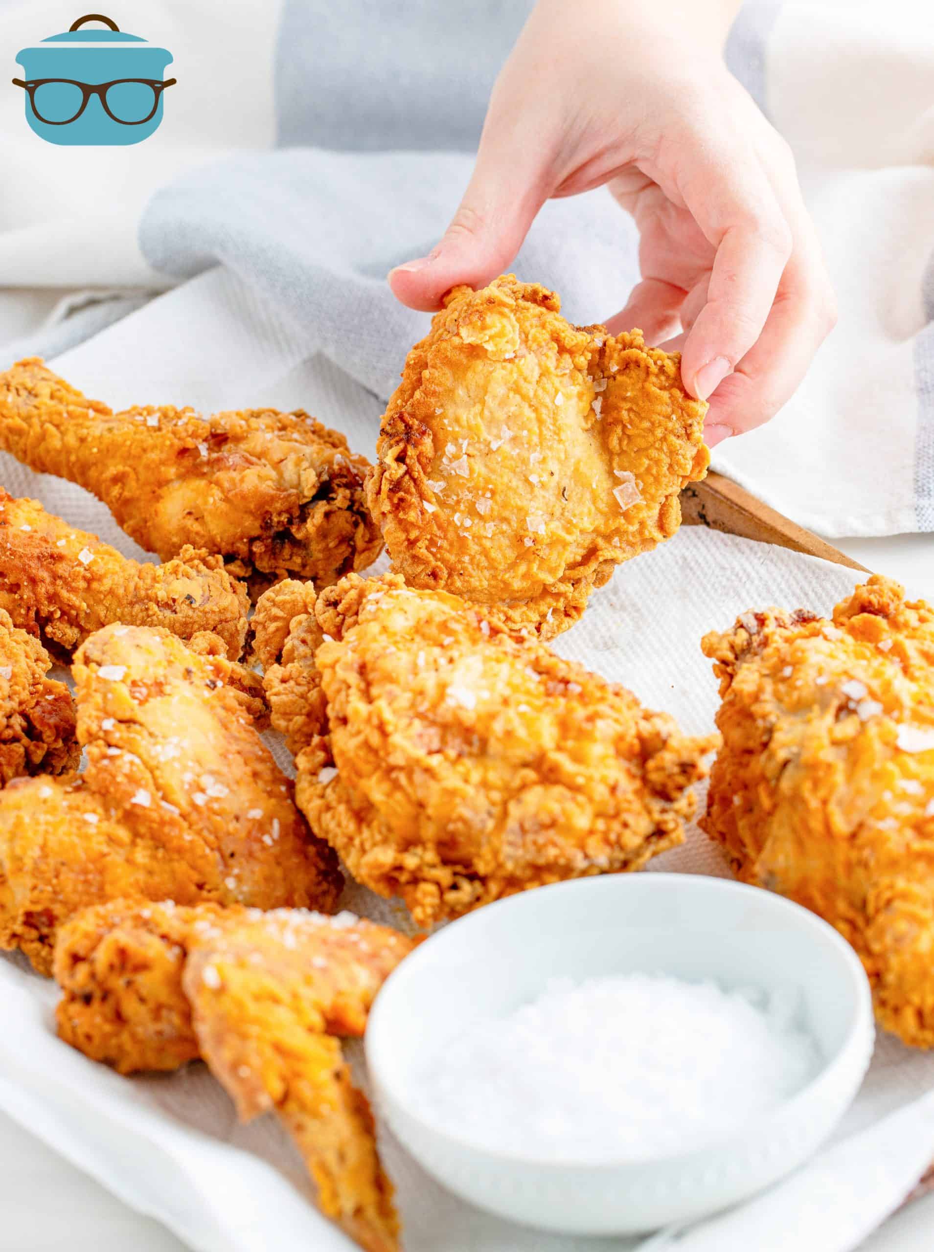 Chicken on paper towels with salt and hand holding up piece of Souther Fried Chicken.