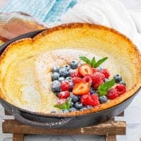 Close up of Dutch Baby Pancake Recipe in skillet topped with powdered sugar and fruit, square image