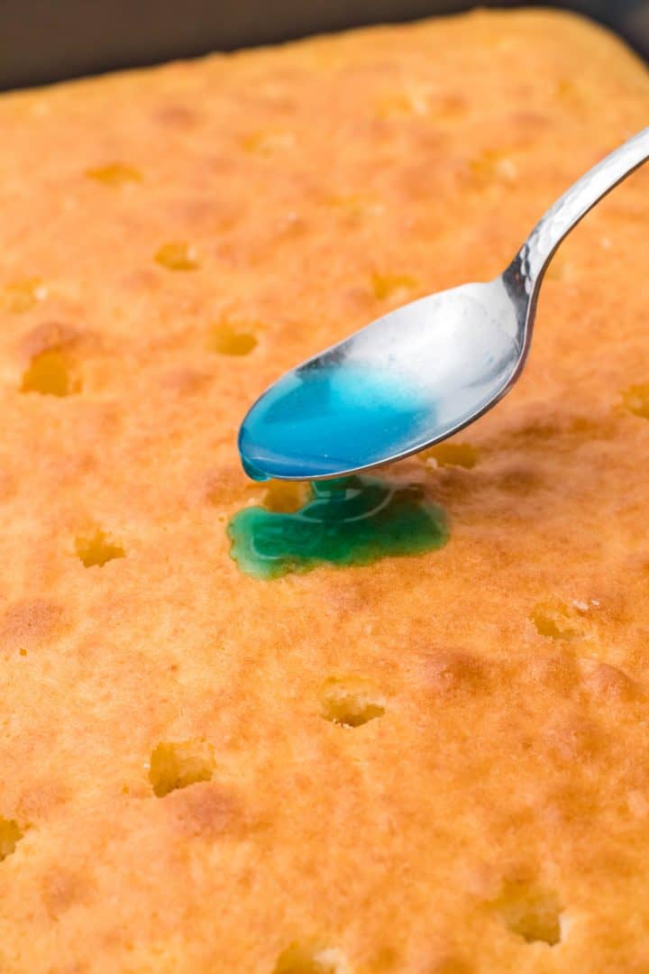 Jello being poured over holes in cake with a spoon.