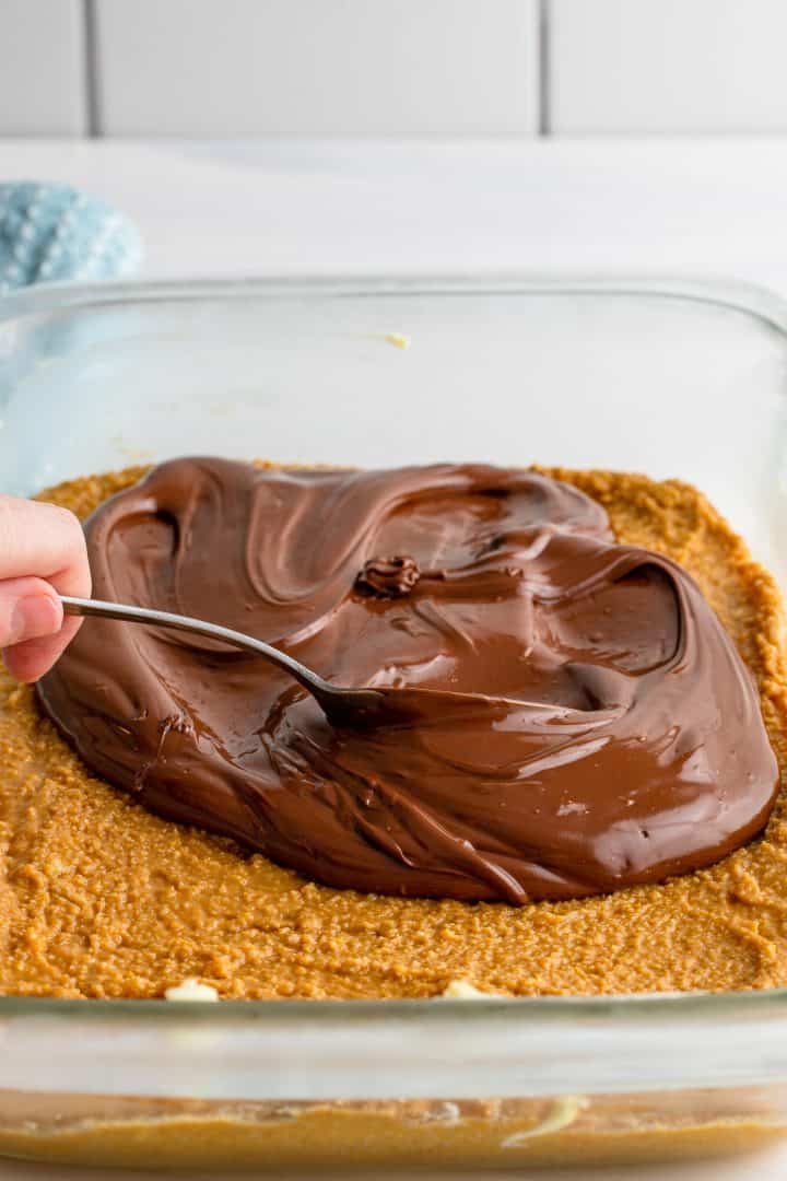 Spoon spreading melted chocolate on top of peanut butter mixture.