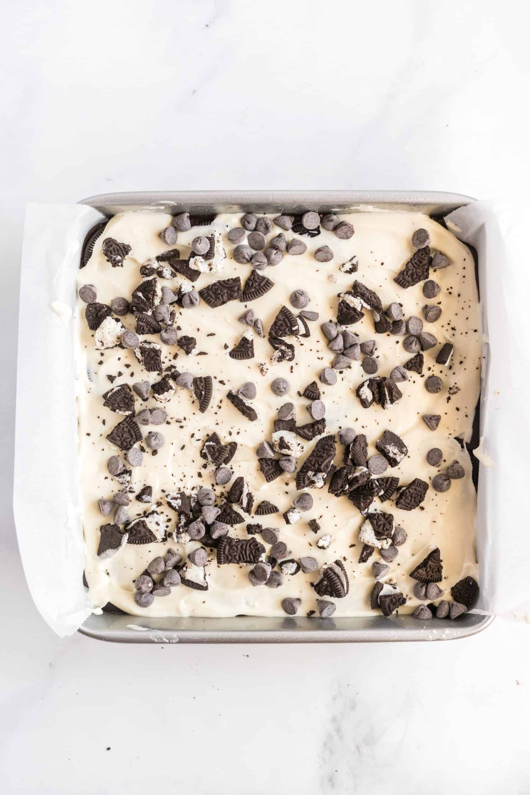 Chopped Oreos and chocolate chips topping cheesecake layer in baking pan.