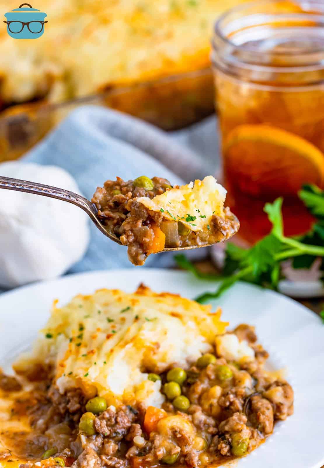 a serving of shepherds pie on a white plate with a fork holding up some of the serving. A glass of iced tea is in the background. 