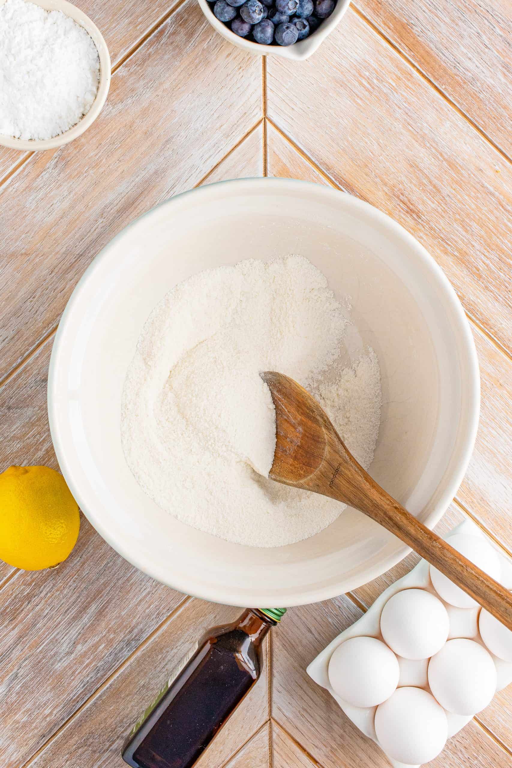 Flour and sugar in white bowl being stirred with wooden spoon.