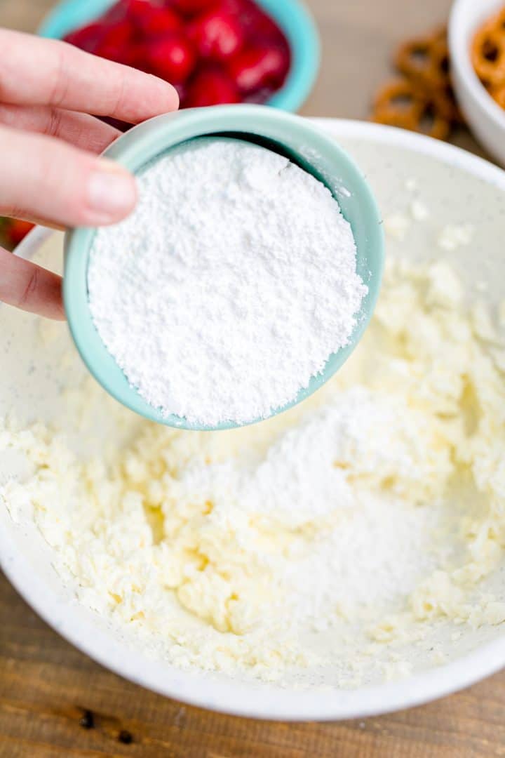Powdered sugar being added to cream cheese in white bowl.