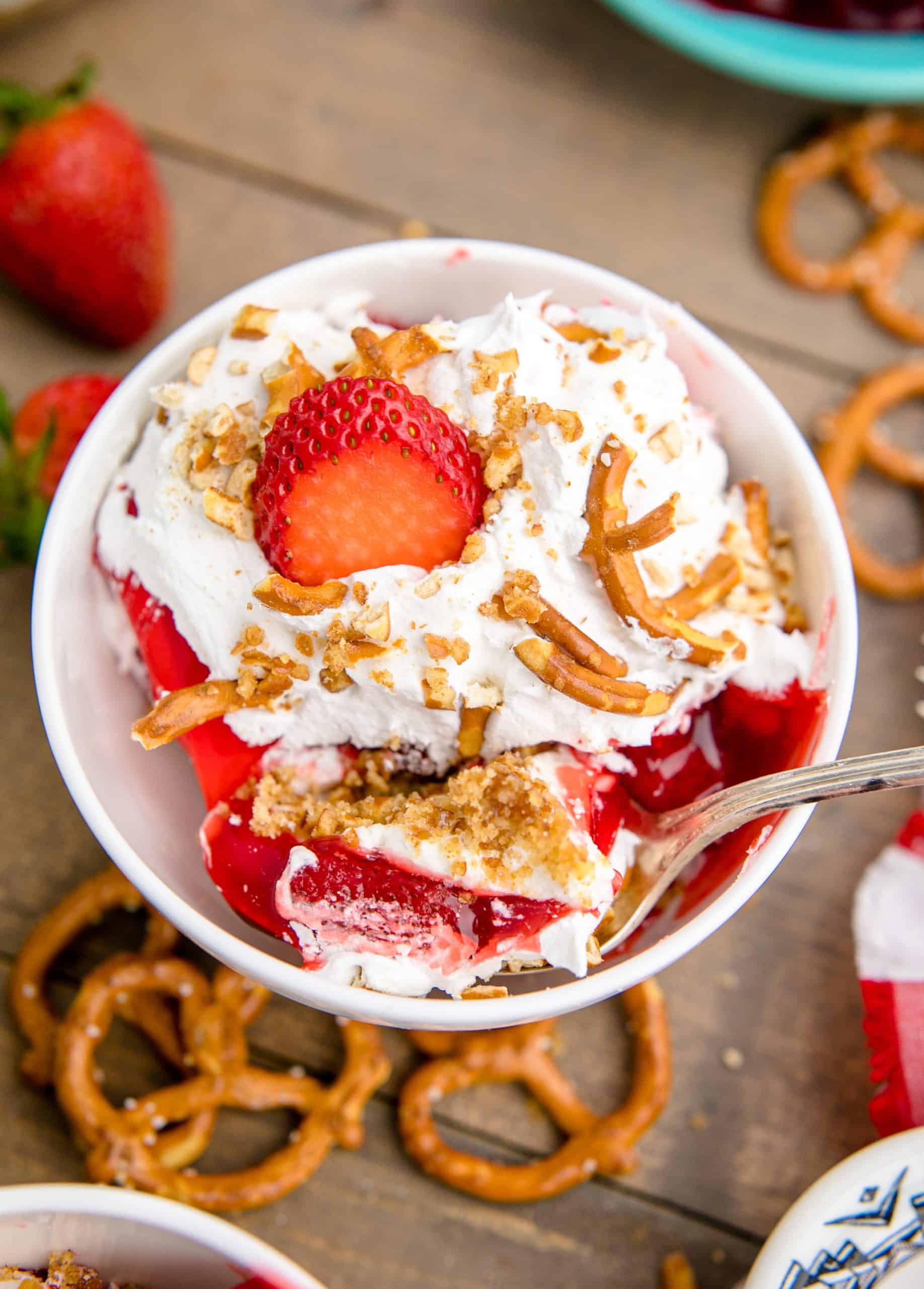 Overhead photo of Strawberry Pretzel Dessert in white bowl with spoon surrounded by pretzels and strawberries.