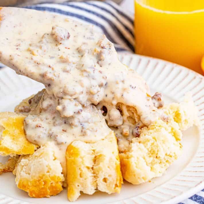 Square up close image of wooden spoon spooning Homemade Sausage Gravy over biscuits on white plate