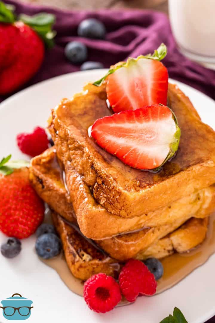 Stacked Brioche French Toast on white plate topped with strawberries and syrup.