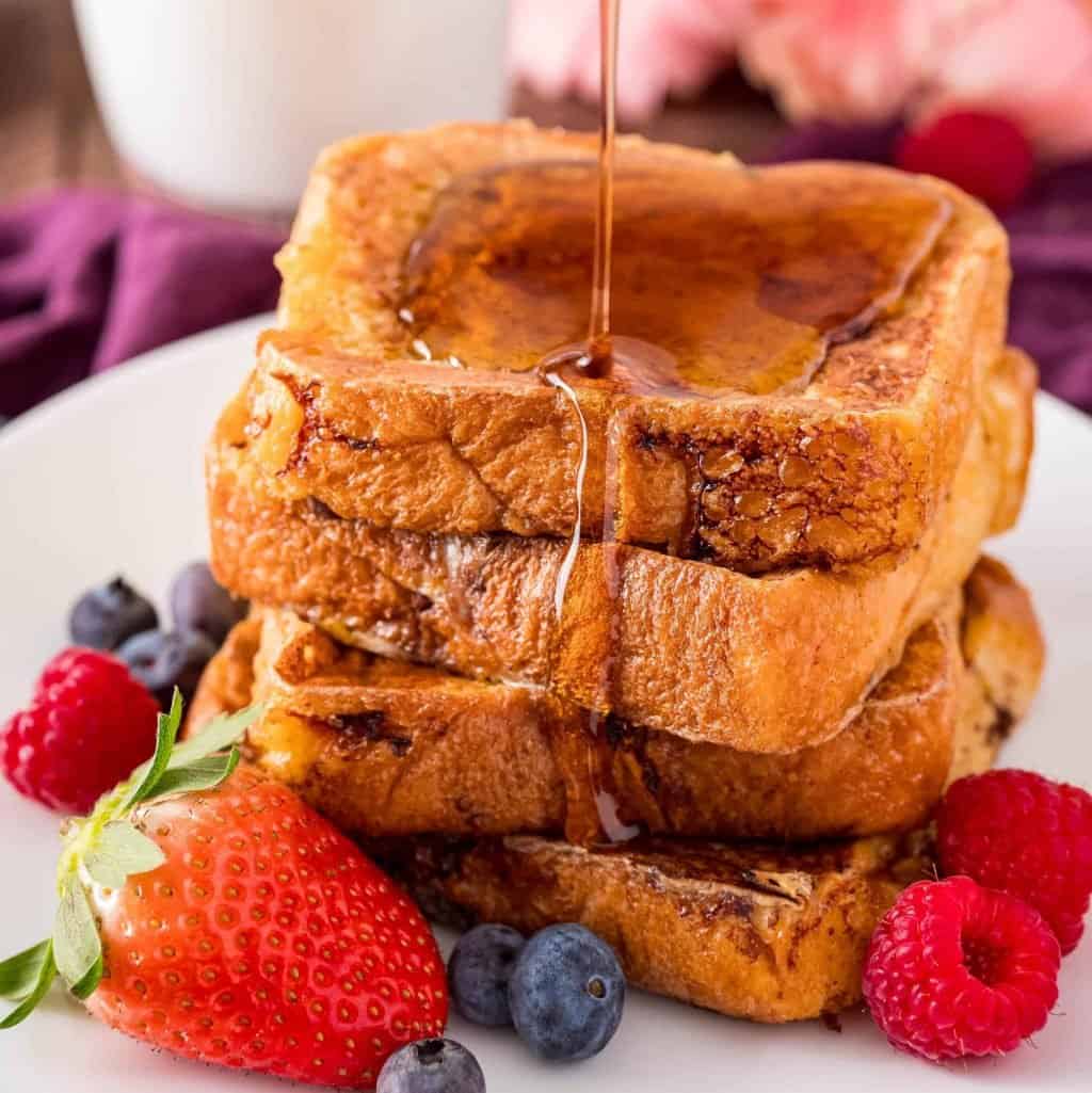 Square photo of syrup being poured over Brioche French Toast on white plate with fruit