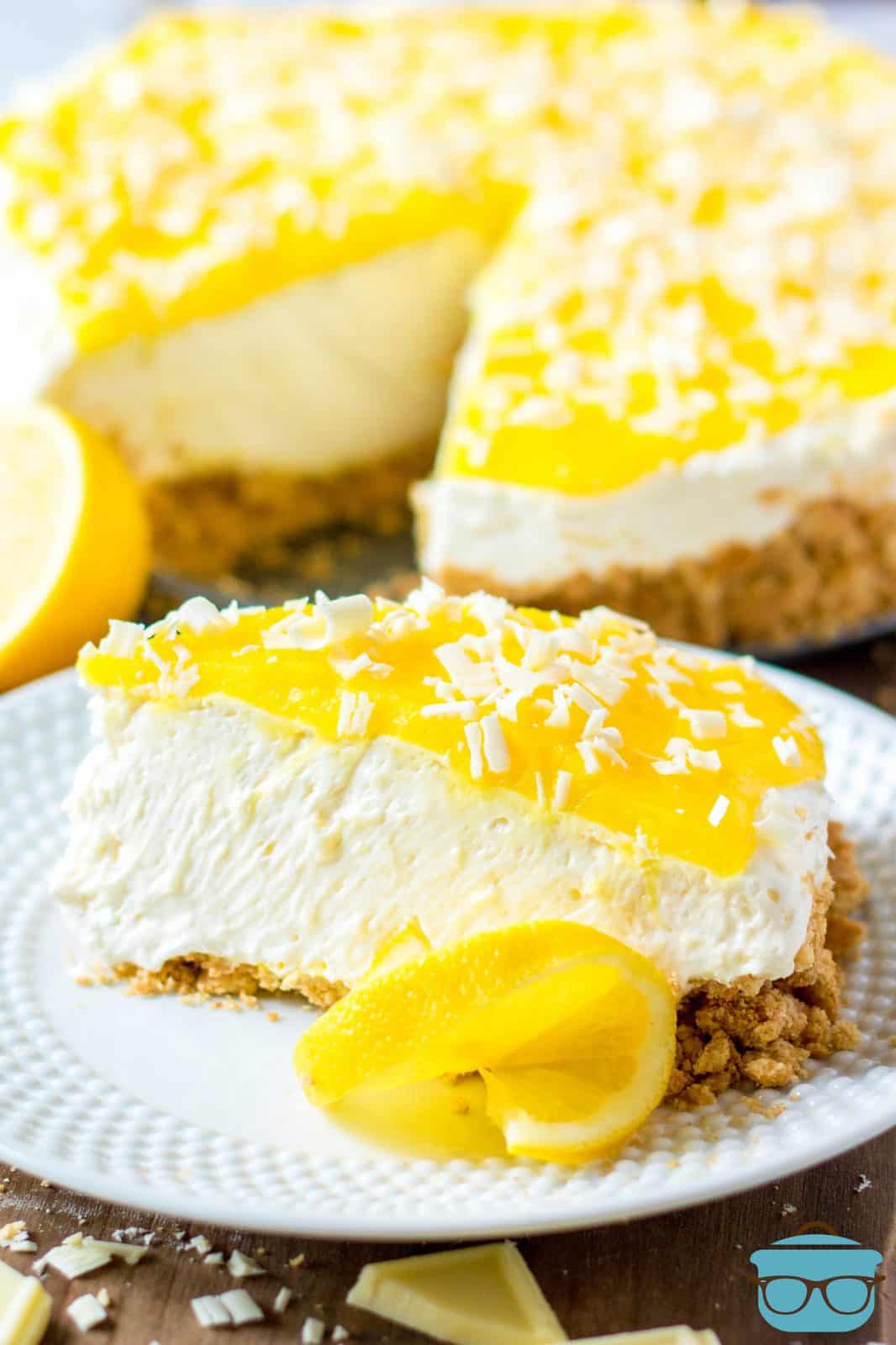 Slice of No-Bake Lemon White Chocolate Cheesecake on white plate with slice of lemon.
