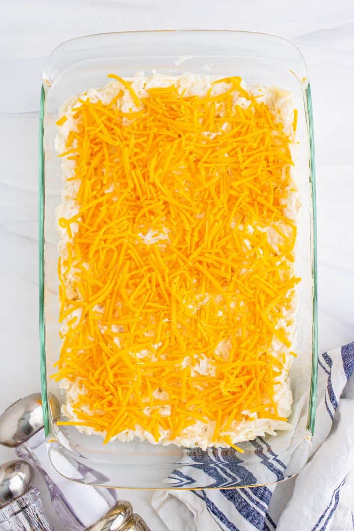 shredded cheese shown sprinkled on hash brown casserole mixture in a glass baking dish