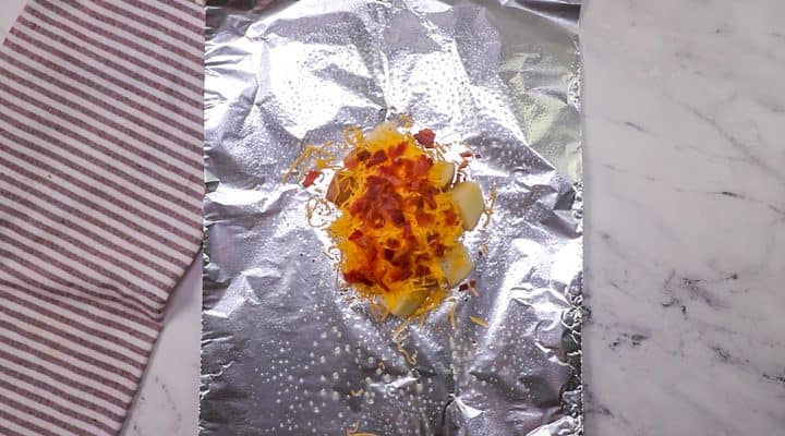 shredded cheese and bacon pieces on top of cubed potatoes and aluminum foil.