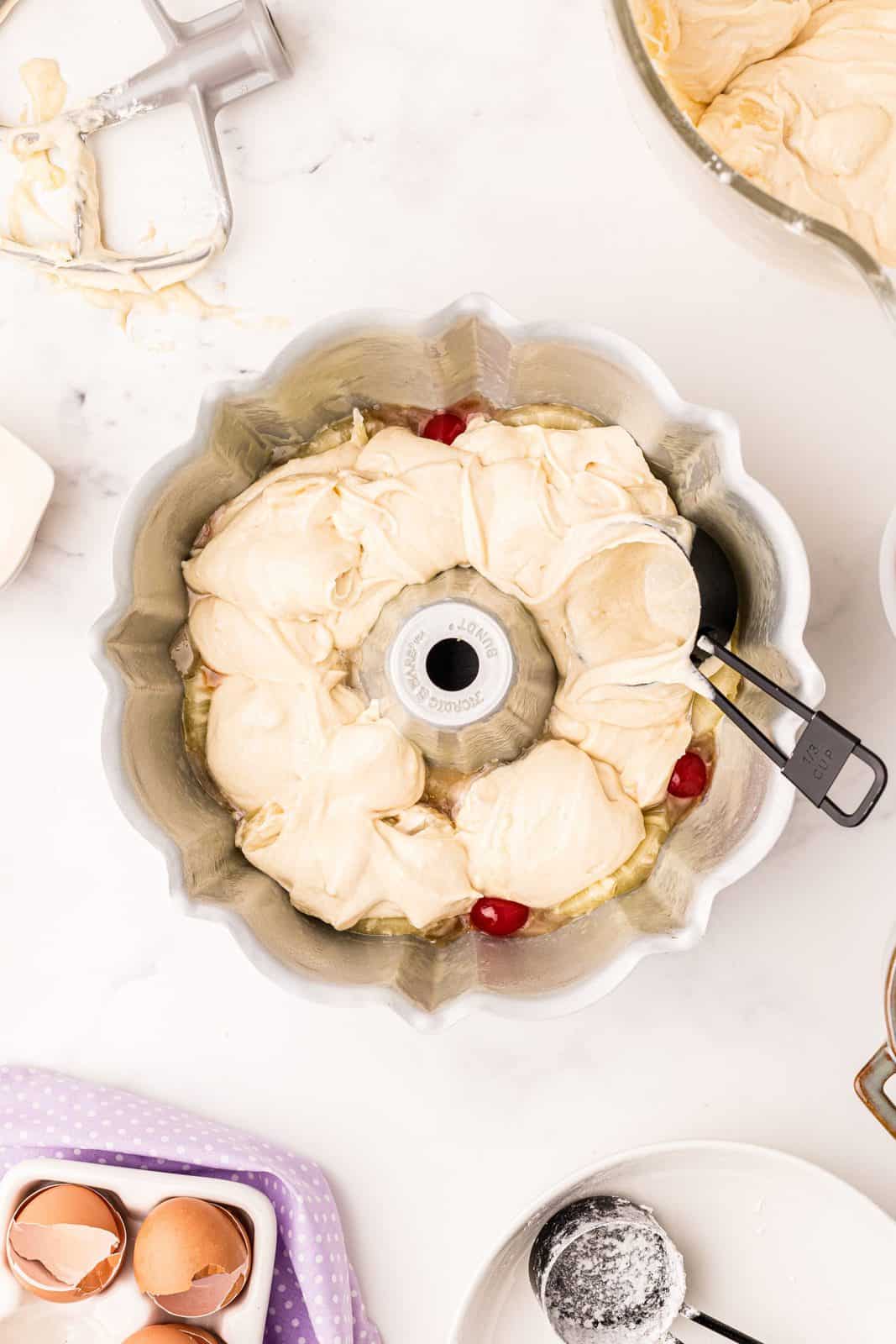 Measuring cup spreading cake batter over pineapple and cherries in bundt pan.
