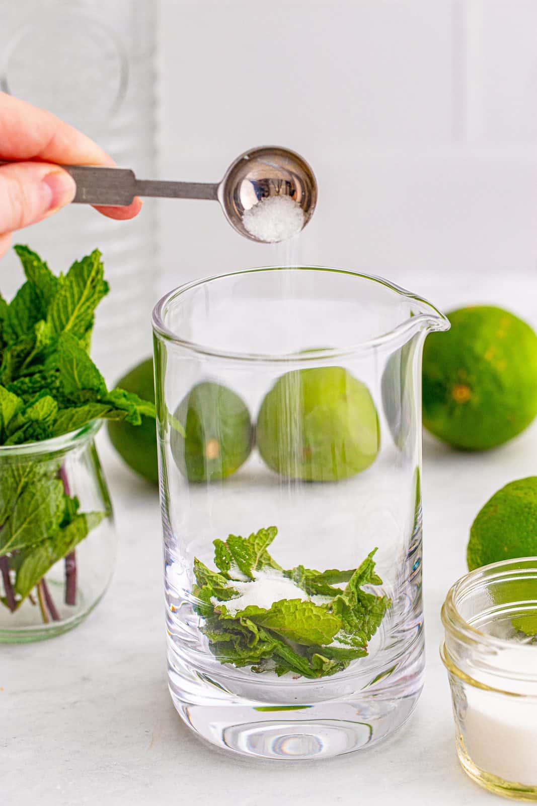 Glass container with mint leaves and sugar being poured in.