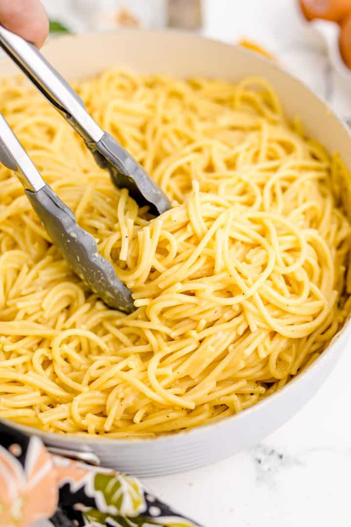 Metal tongs tossing spaghetti carbonara mixture together in skillet.