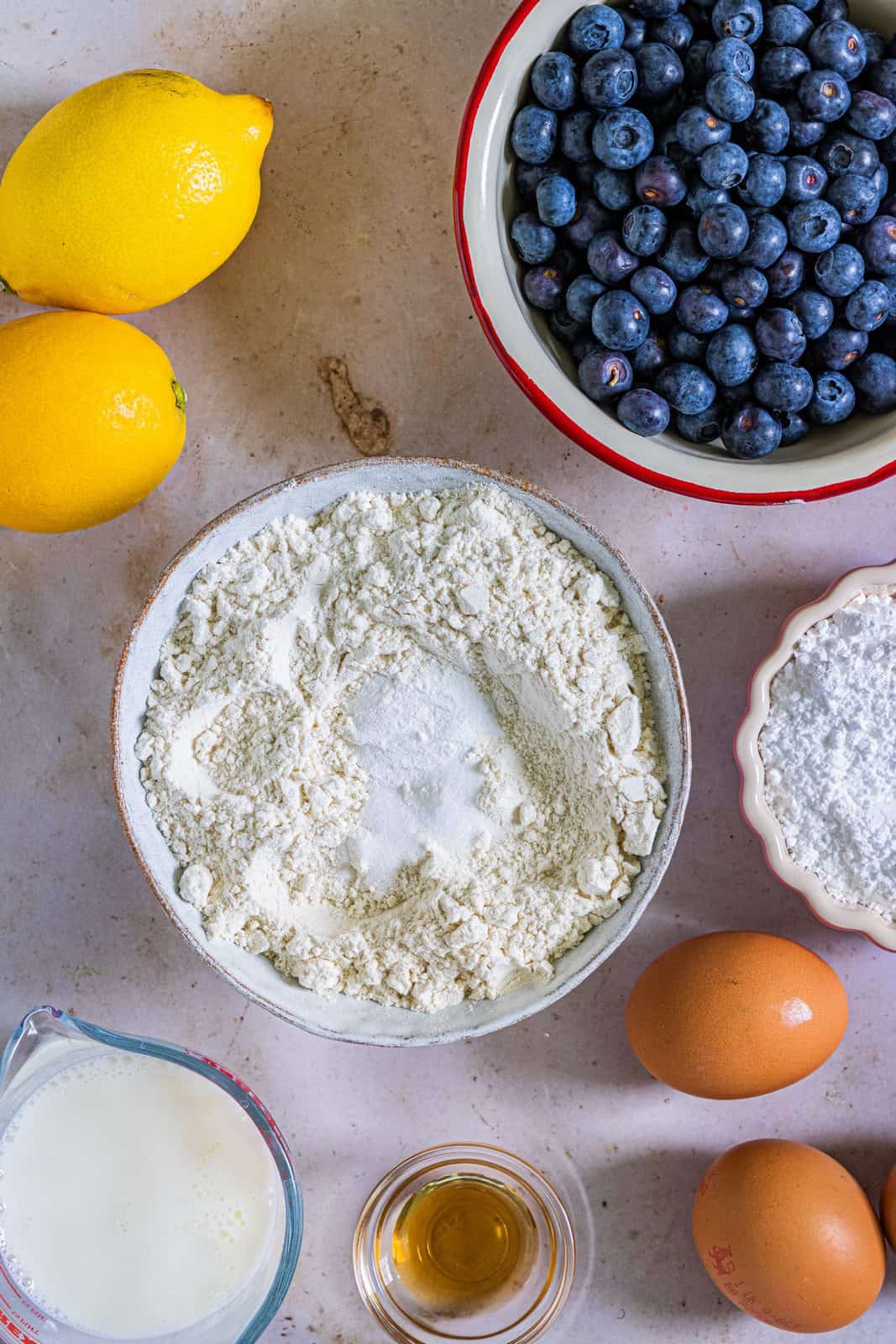 Dry ingredients in white bowl surrounded by fresh blueberries, lemon, eggs and milk.