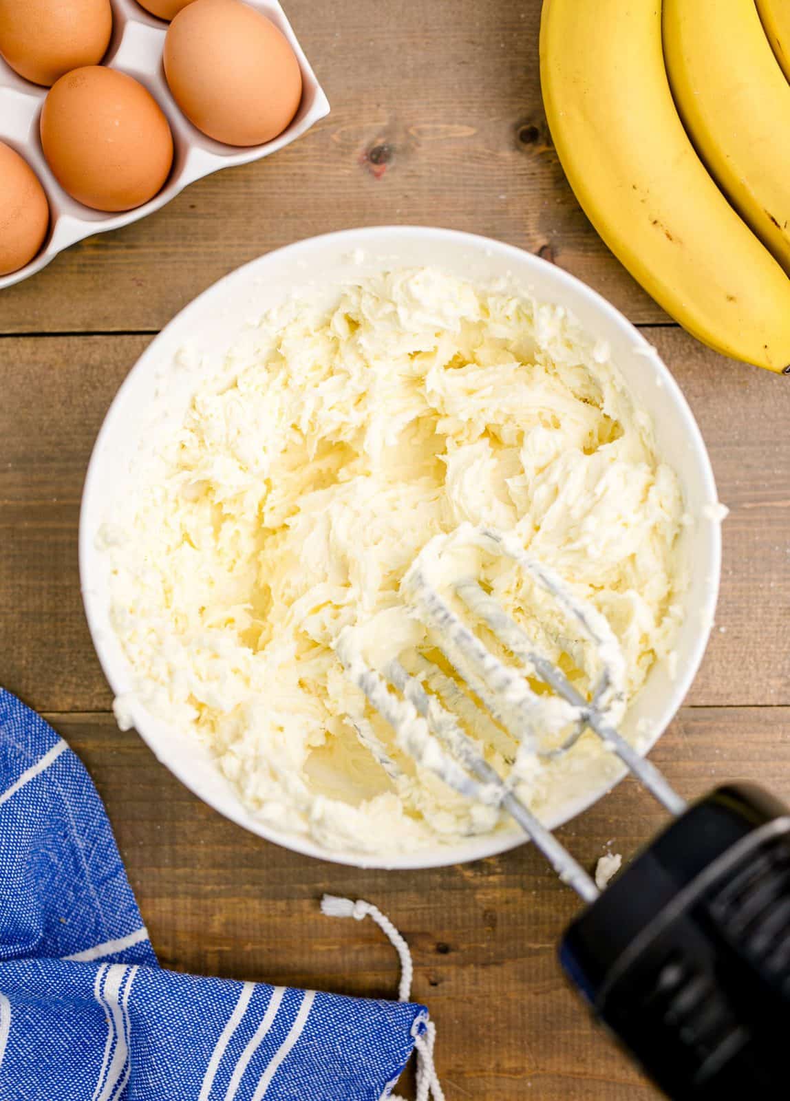 Cream cheese and sugar blended together in white bowl with a handheld electric mixer.