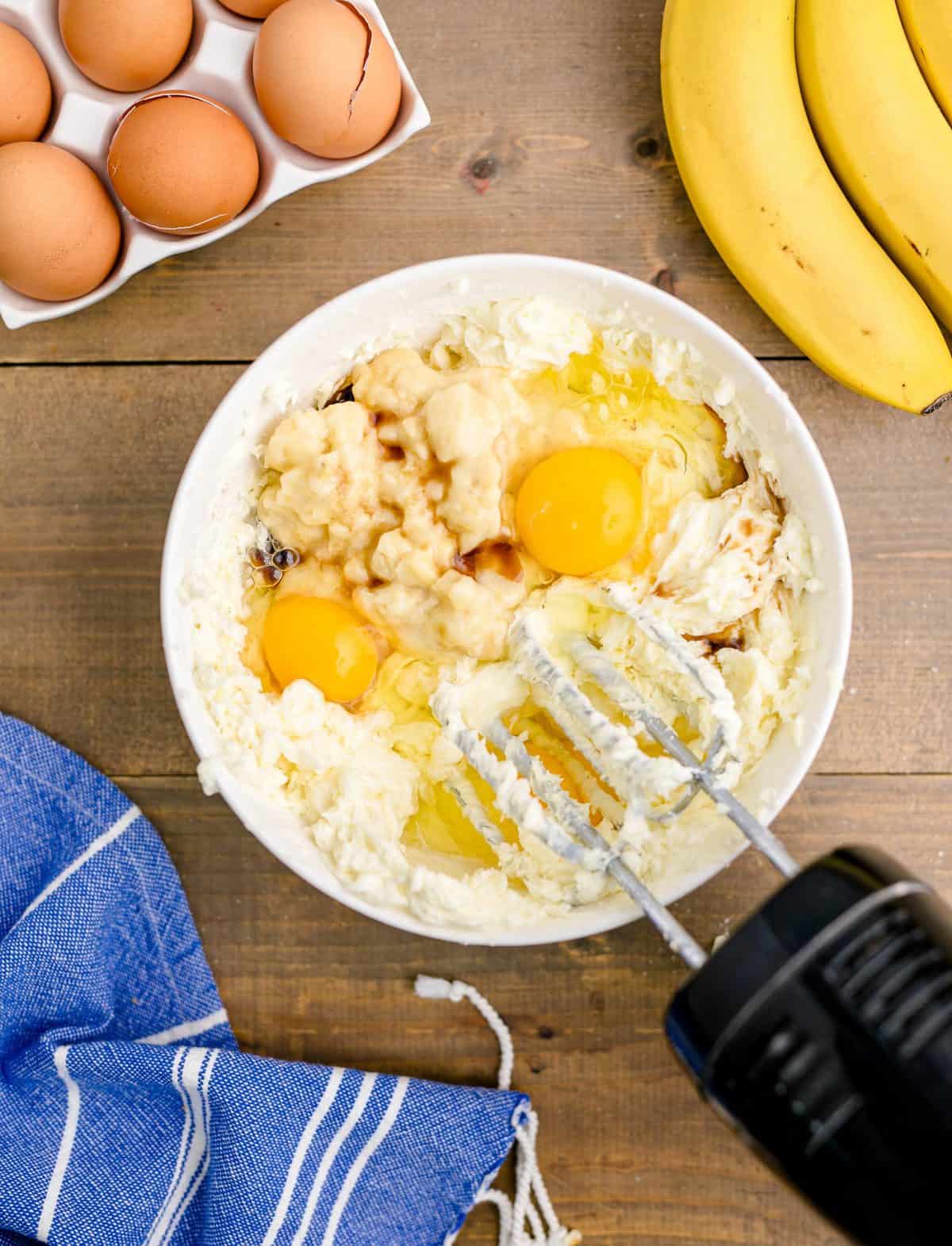 Eggs, vanilla and banana added to cream cheese mixture in a white bowl.