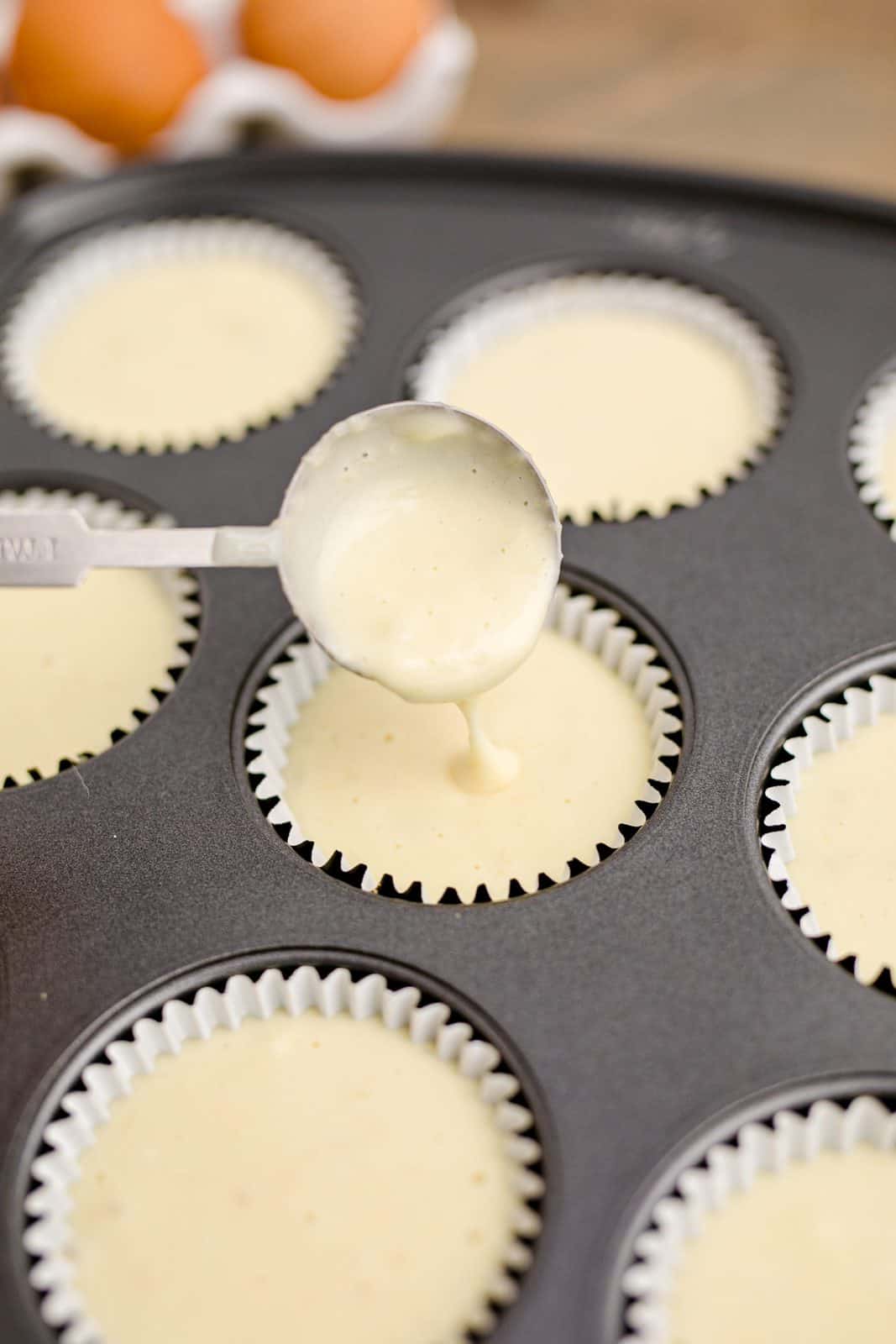 Cheesecake filling being spooned into paper liners in muffin tin.