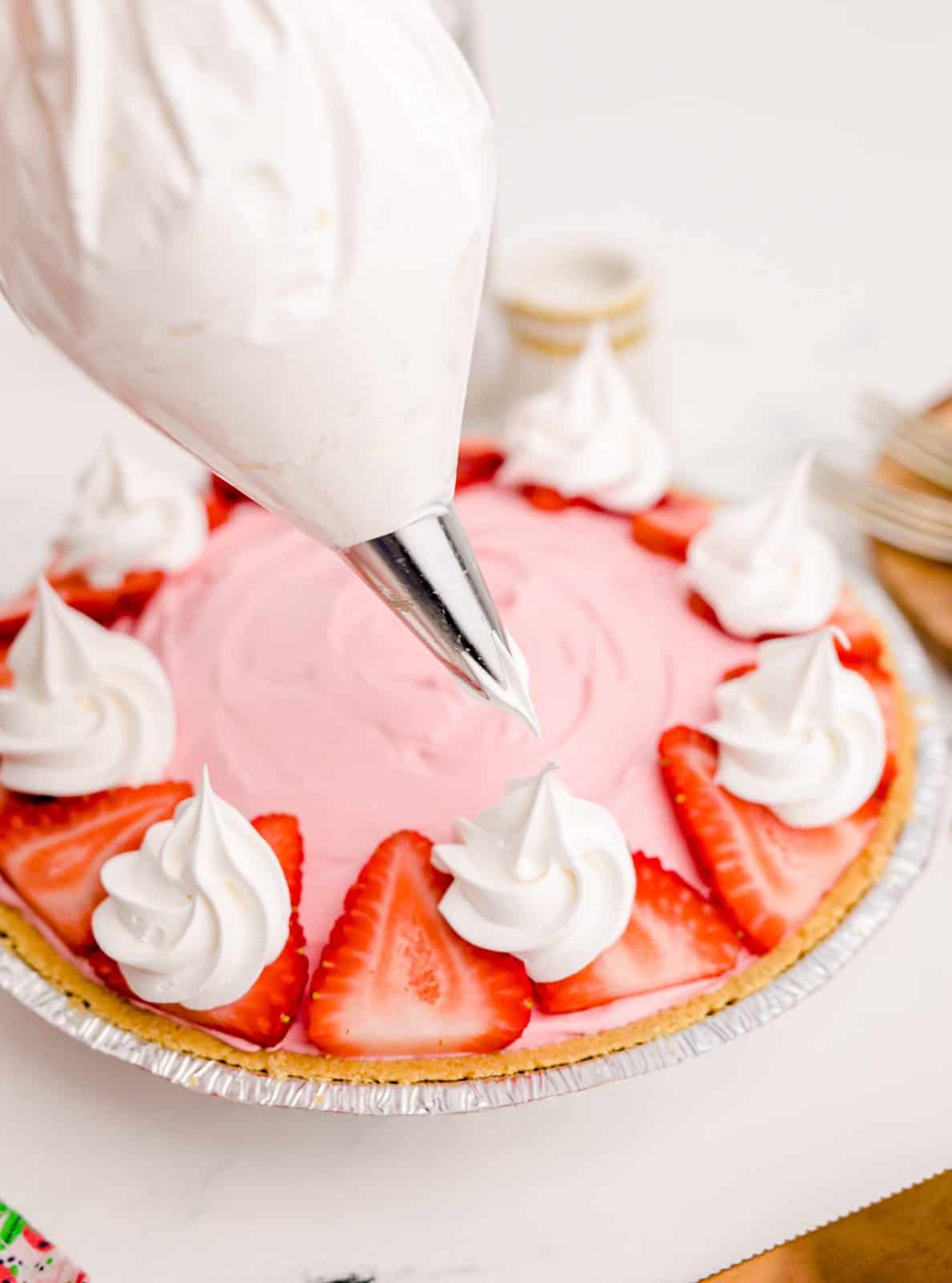Strawberry Kool-Aid Pie Recipe being garnished with strawberries and cool whip. A piping bag is being held over the pie.