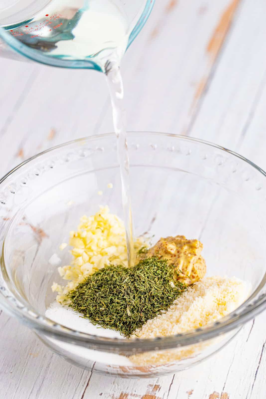 white wine vinegar being poured in a bowl that has sugar, mustard, minced garlic, dill and Parmesan cheese in it. 