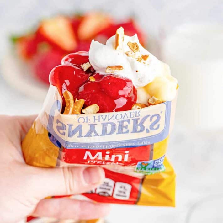 Square image of hand holding one bag of Walking Strawberry Pretzel Salad Recipe showing ingredients poking out of top.
