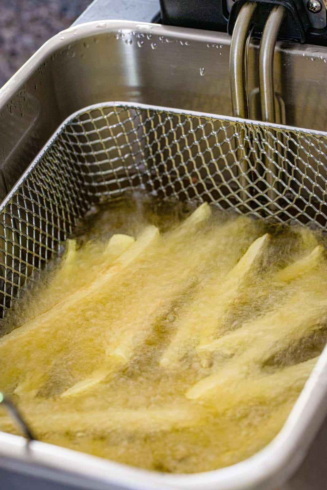 sliced potatoes shown bubbling in oil in a deep fryer