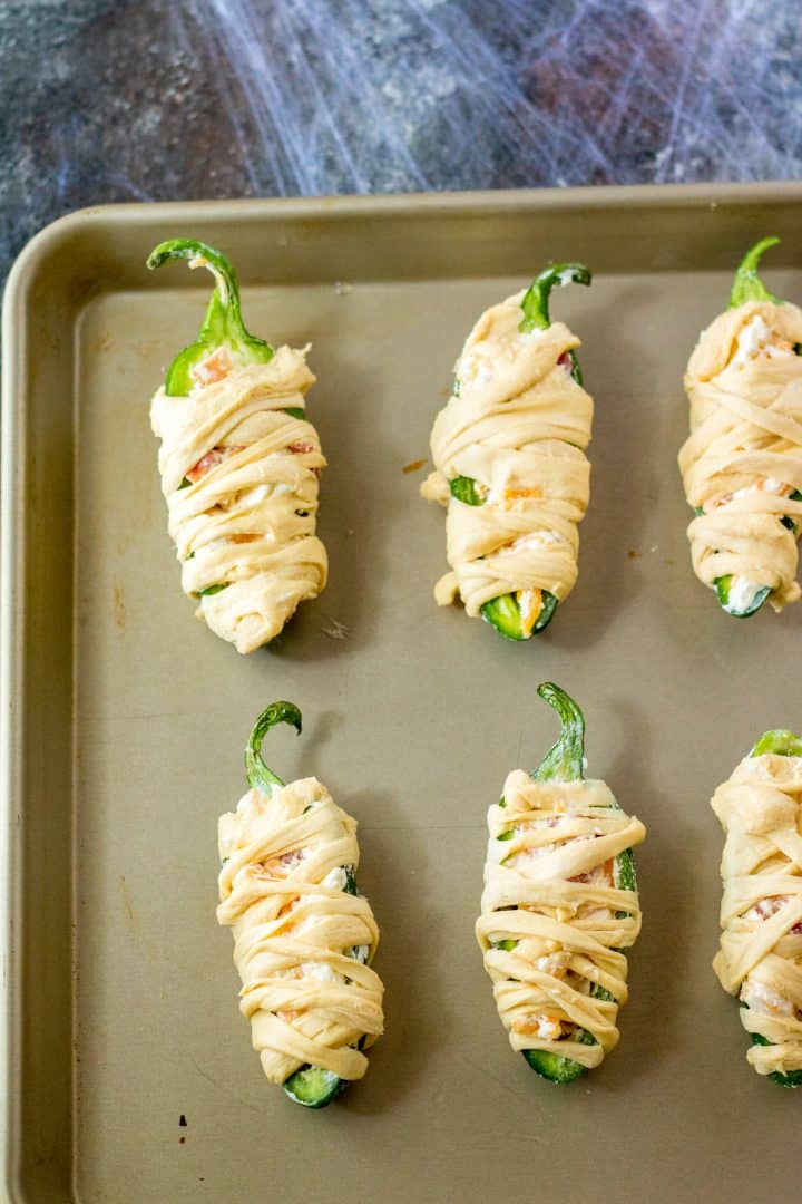 Jalapenos wrapped in strips of crescent rolls on baking sheet.