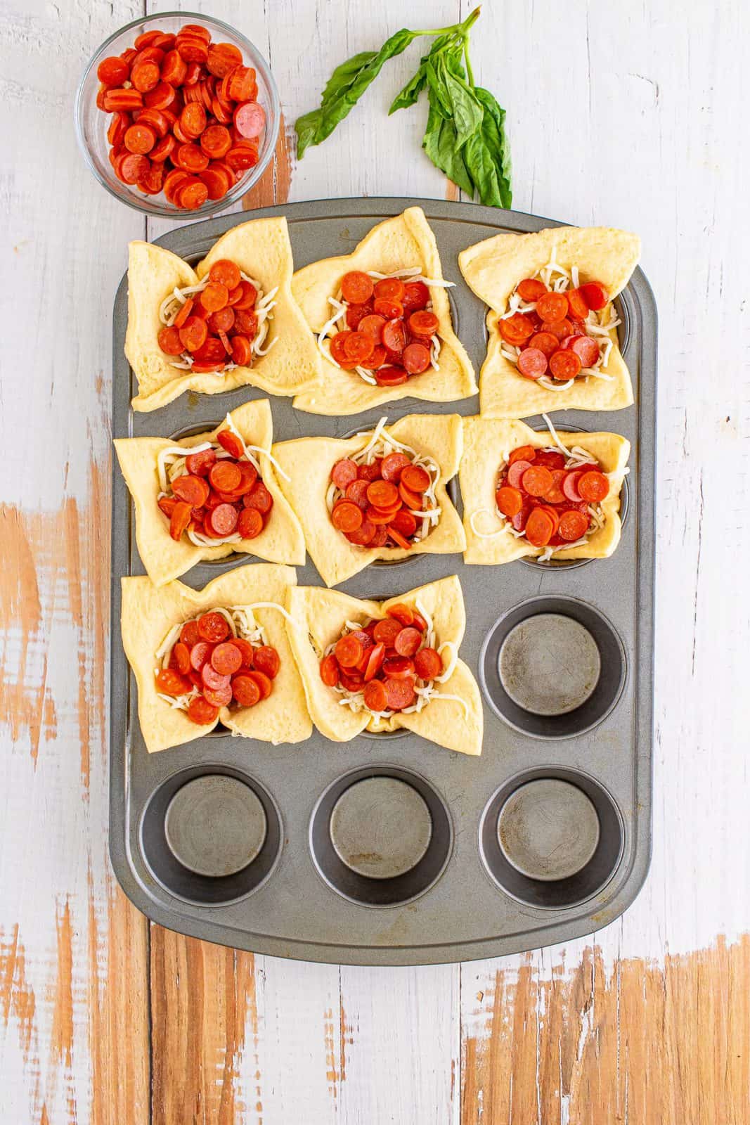 Mini pepperoni placed on top of pizza sauce in muffin tin.