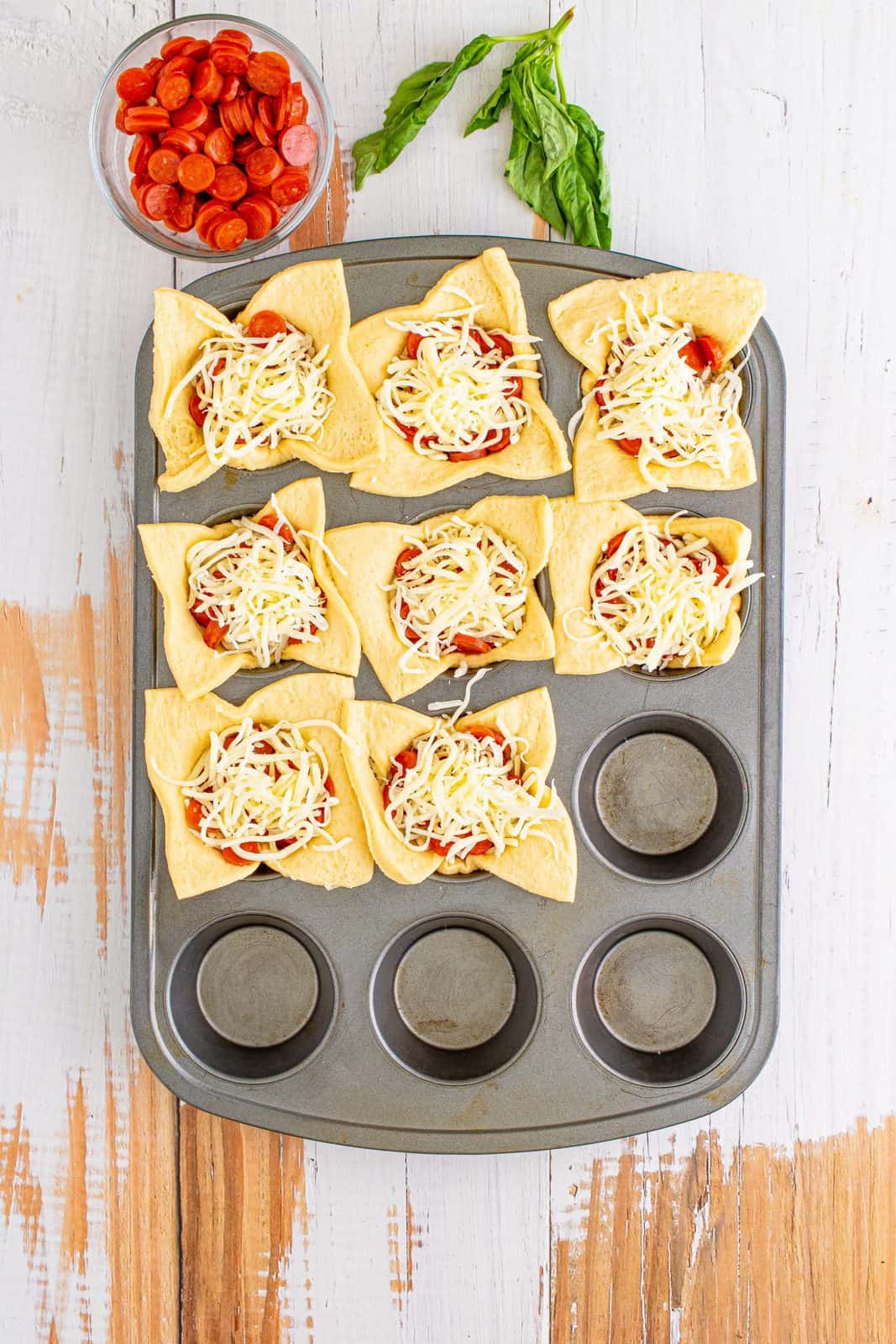 Cheese sprinkled over tops of pepperoni in muffin tin.
