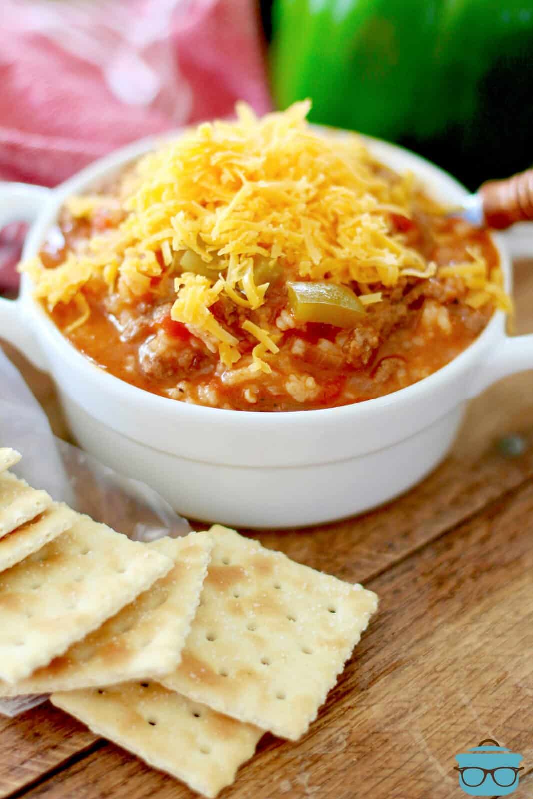 stuffed pepper soup shown in a small white bowl with handles and topped with shredded cheddar cheese and crackers on the side. 