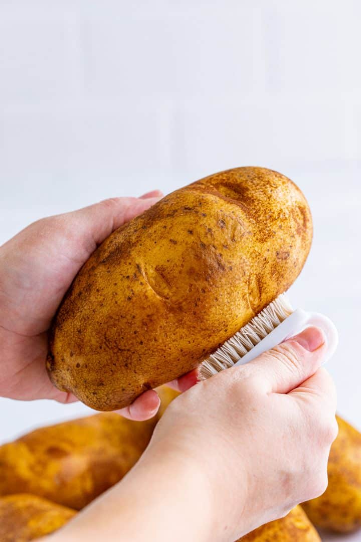 Hands scrubbing potato clean with a vegetable scrubber.