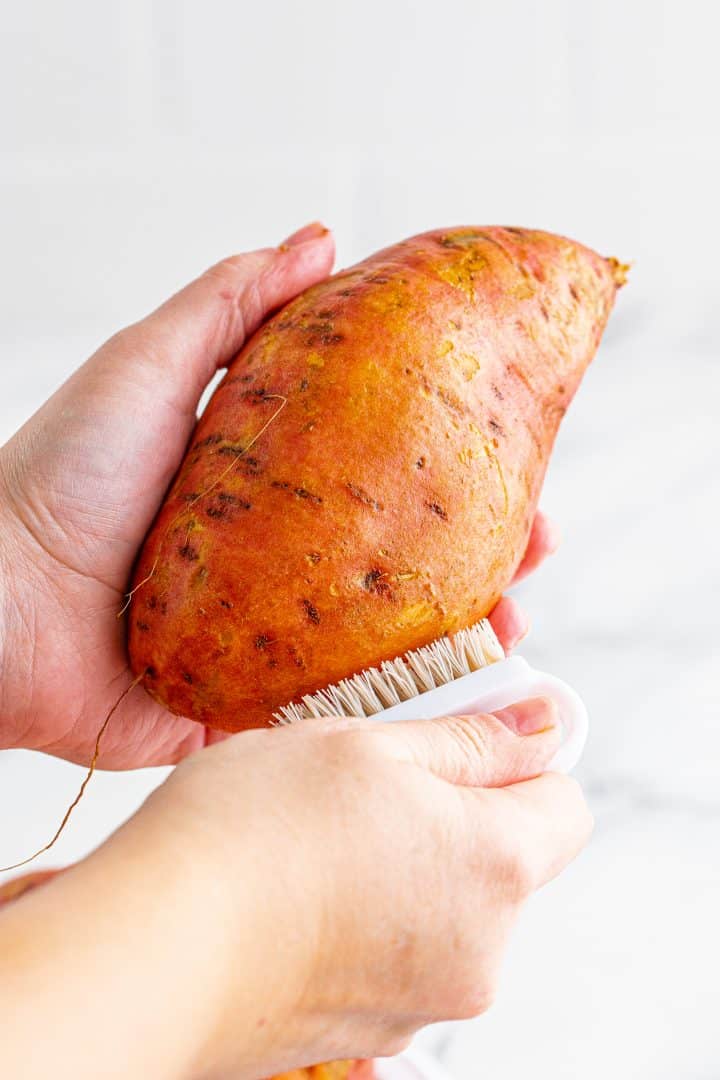 Hands scrubbing sweet potatoes with a vegetable scrubber.