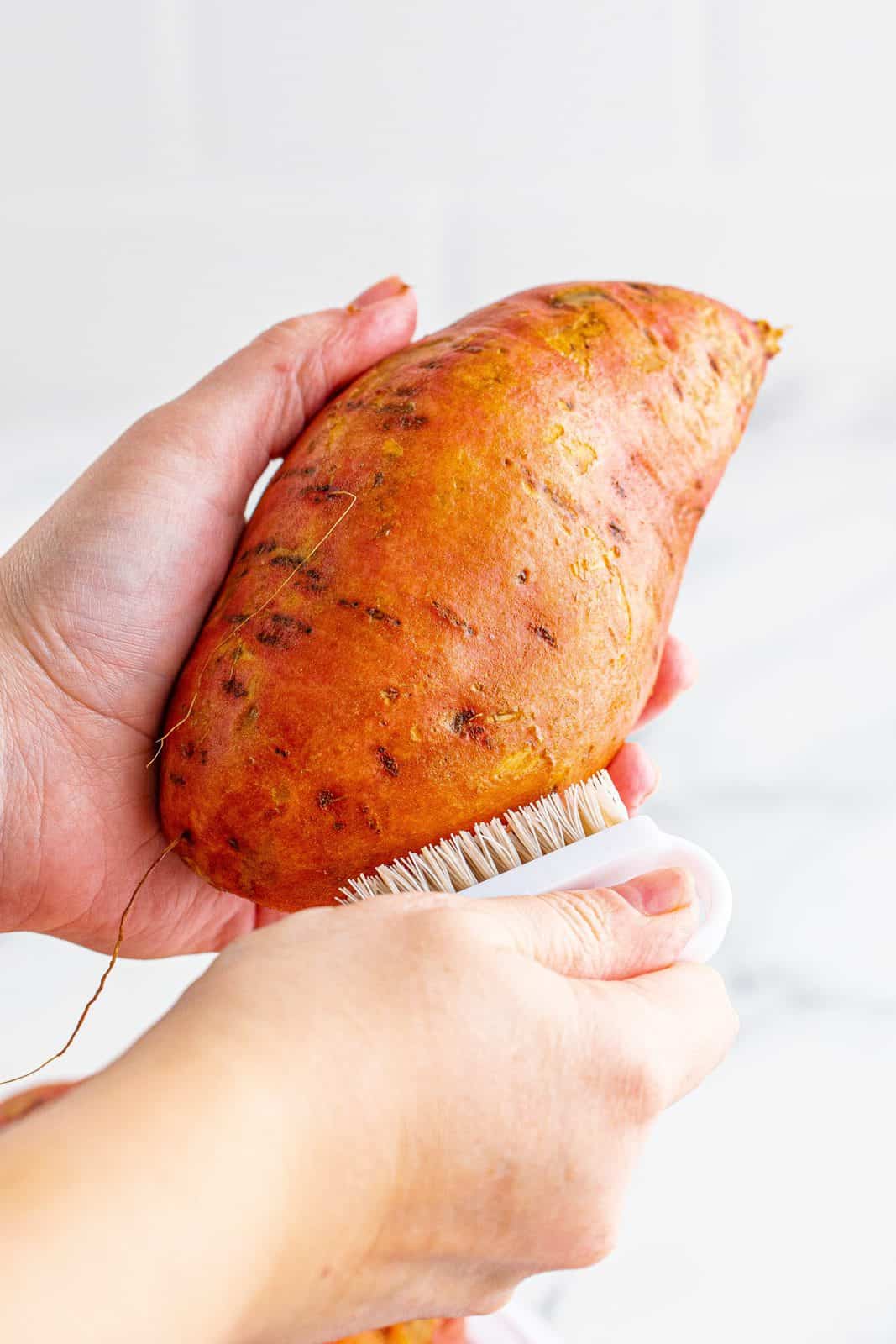 Hands scrubbing sweet potatoes with a vegetable scrubber.