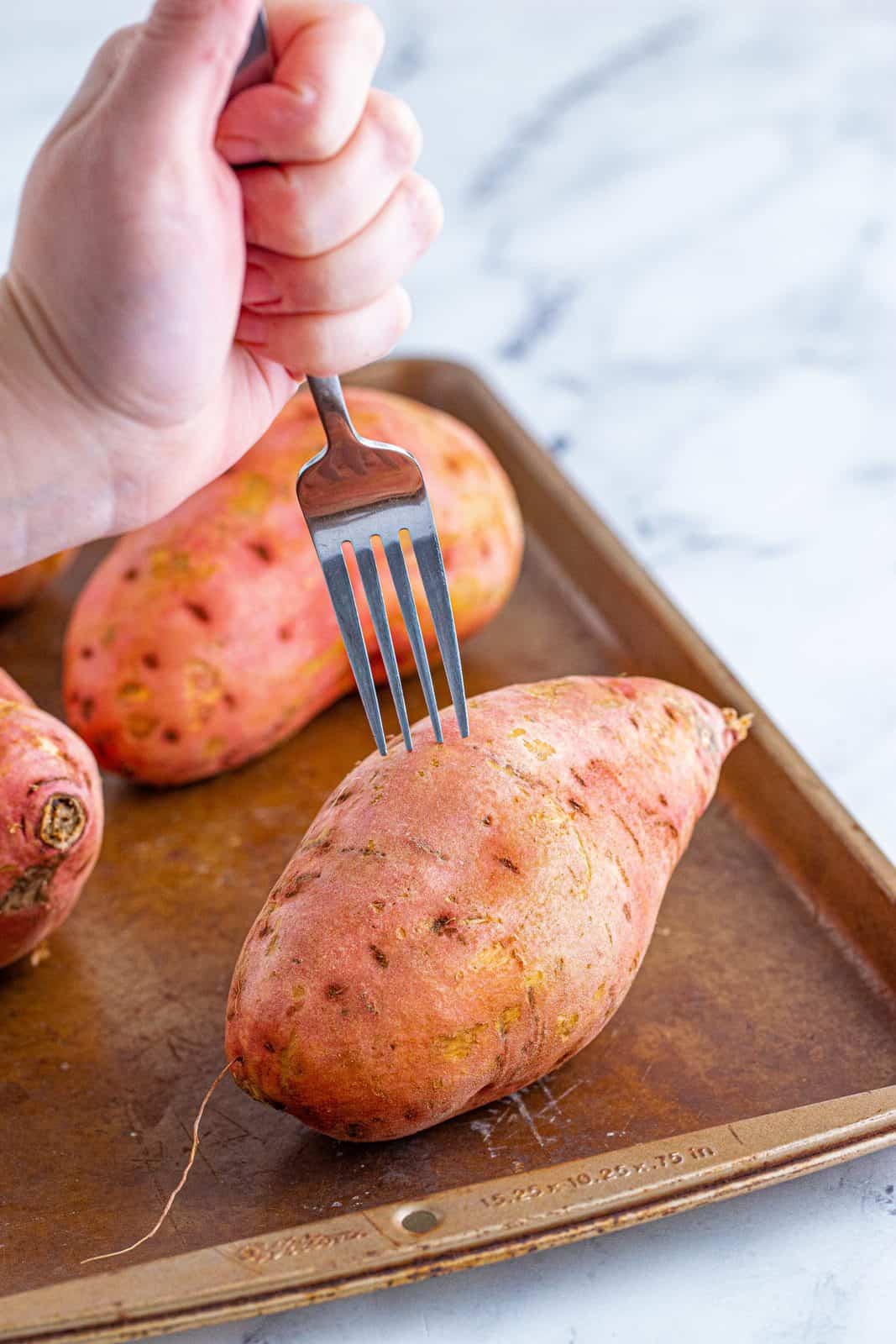 Sweet potatoes on baking pan being pierced with fork.