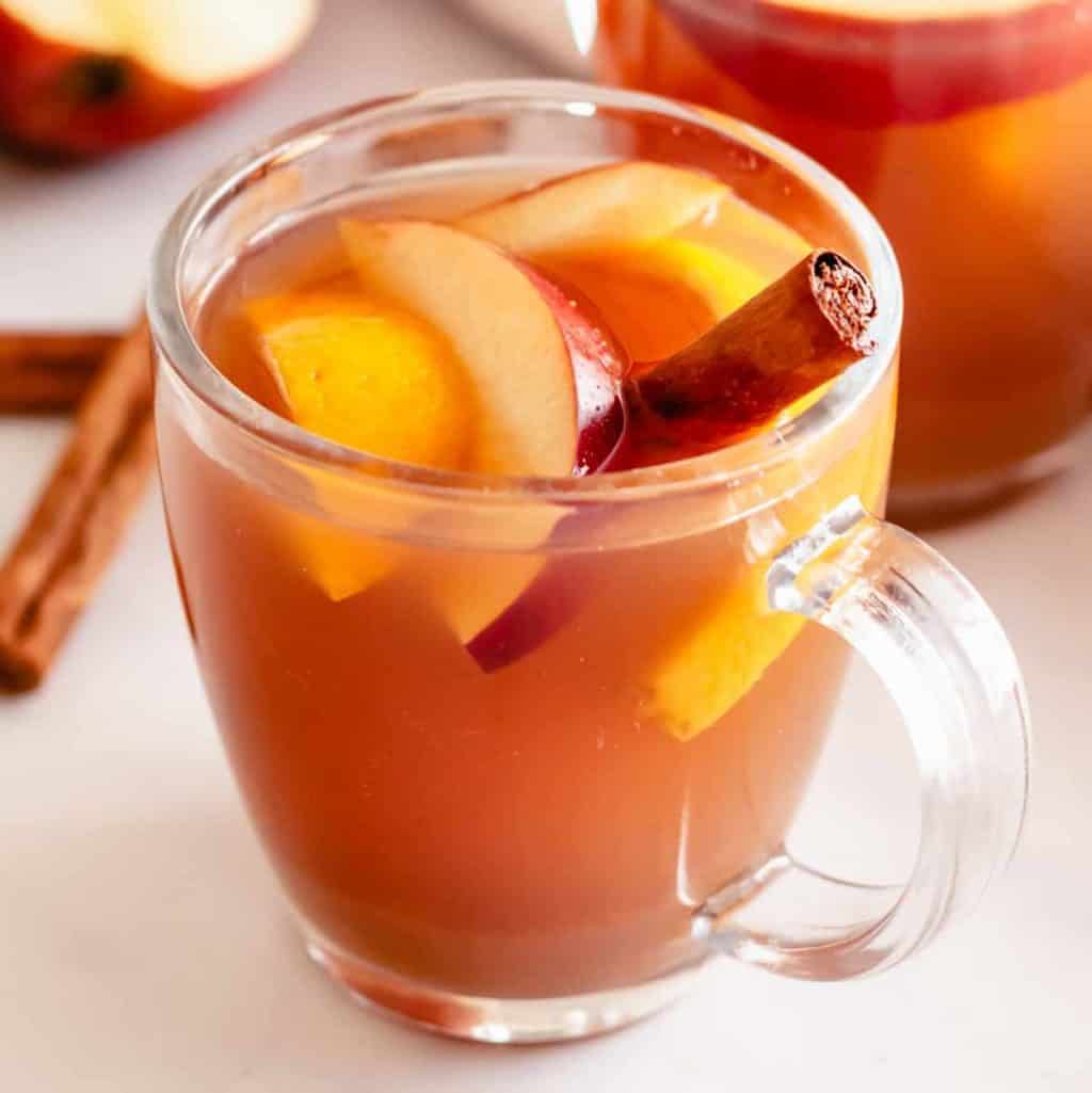 Square image of finished Homemade Apple Cider in clear mug with apples and cinnamon stick.