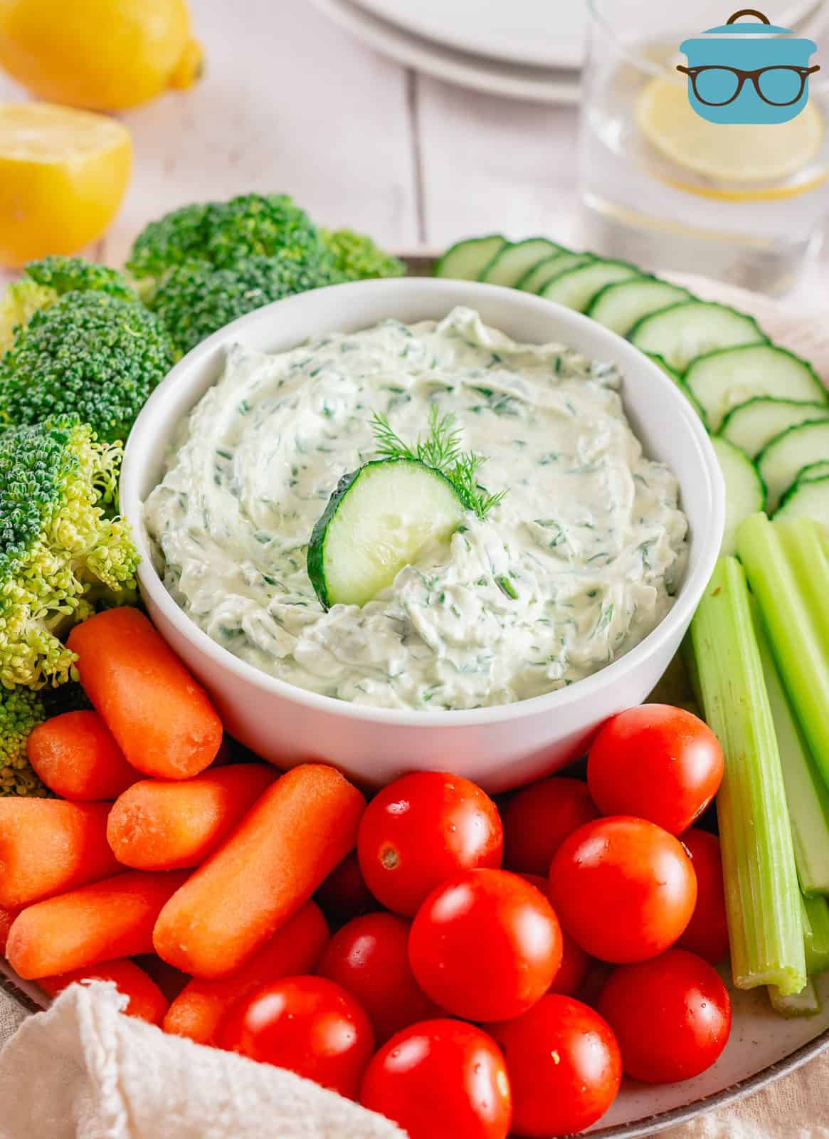 Spinach Dip in bowl surrounded by vegetables and slice of cucumber in bowl.